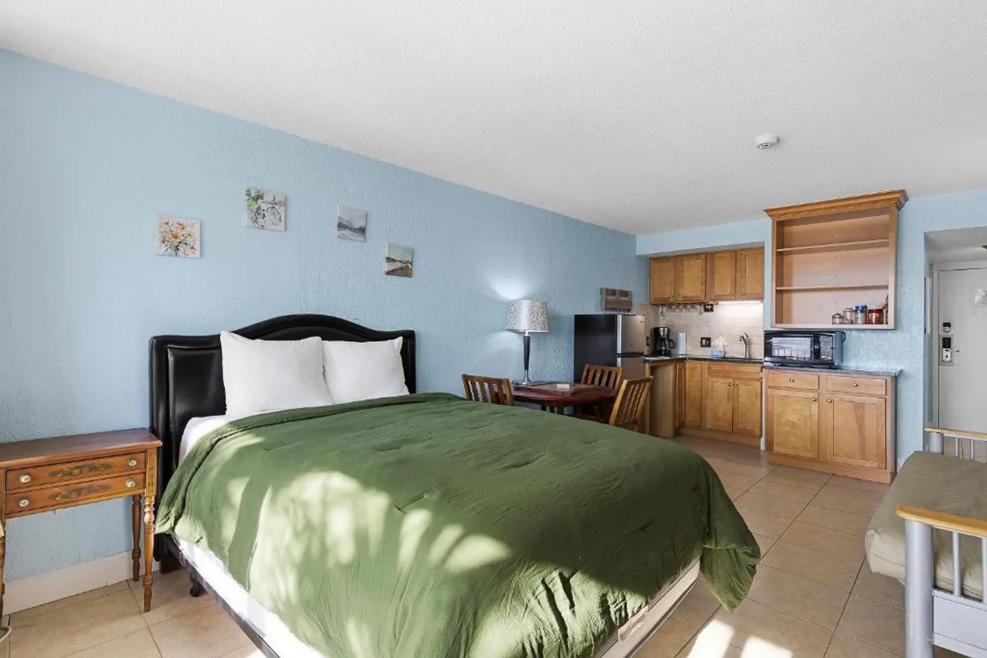 Studio apartment interior showing a bed with a green comforter, dining table, and a small kitchenette with wooden cabinets.