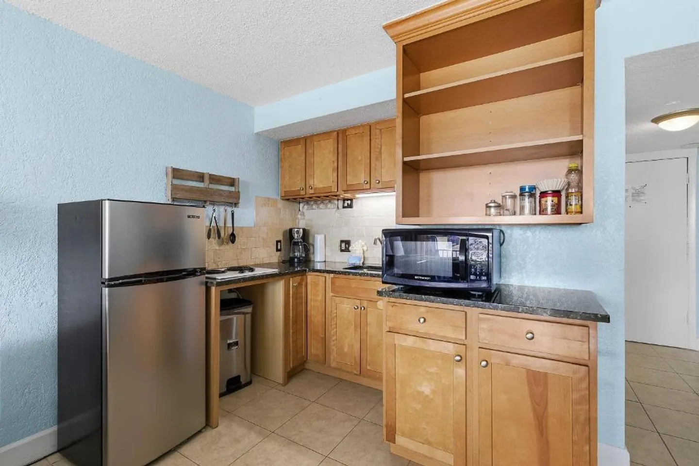 Compact kitchen area with light wooden cabinets, light blue textured walls, small refrigerator, and microwave.