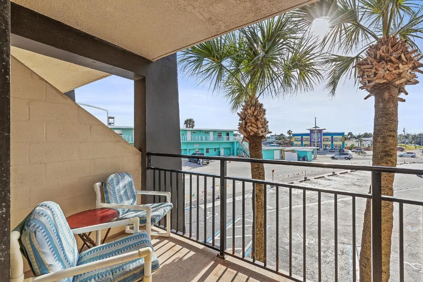 Balcony with two striped armchairs and a small table overlooking palm trees and a parking area under bright sunlight.