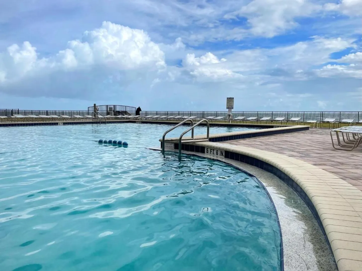 Large outdoor swimming pool with a paved deck, lounge chairs, and safety railings under a blue, partly cloudy sky.