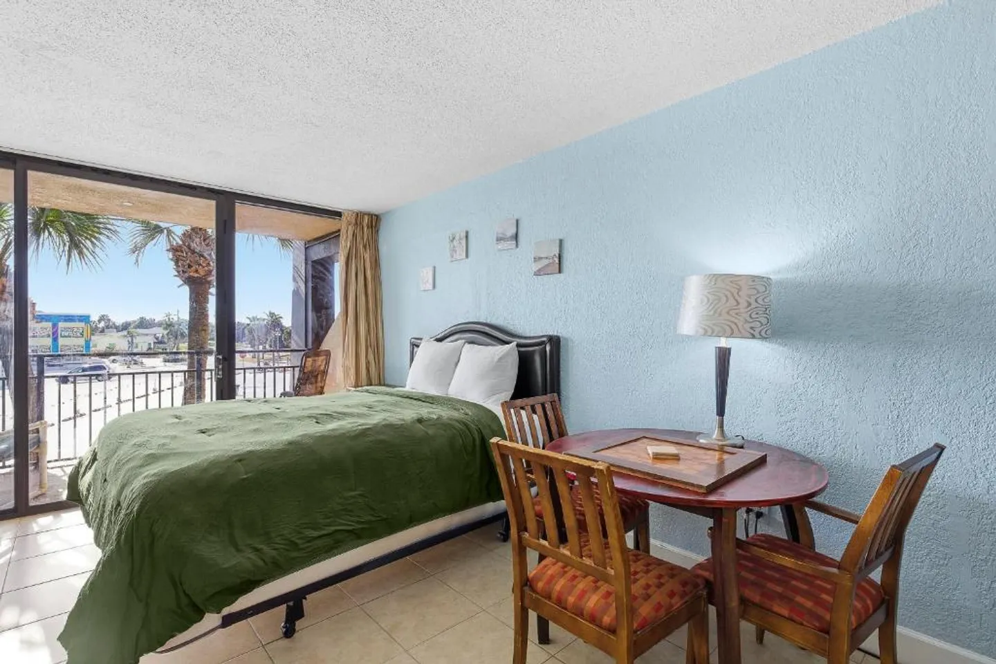 Bedroom with a bed covered in a green blanket next to sliding glass doors, a small dining table with chairs, and light blue textured walls.