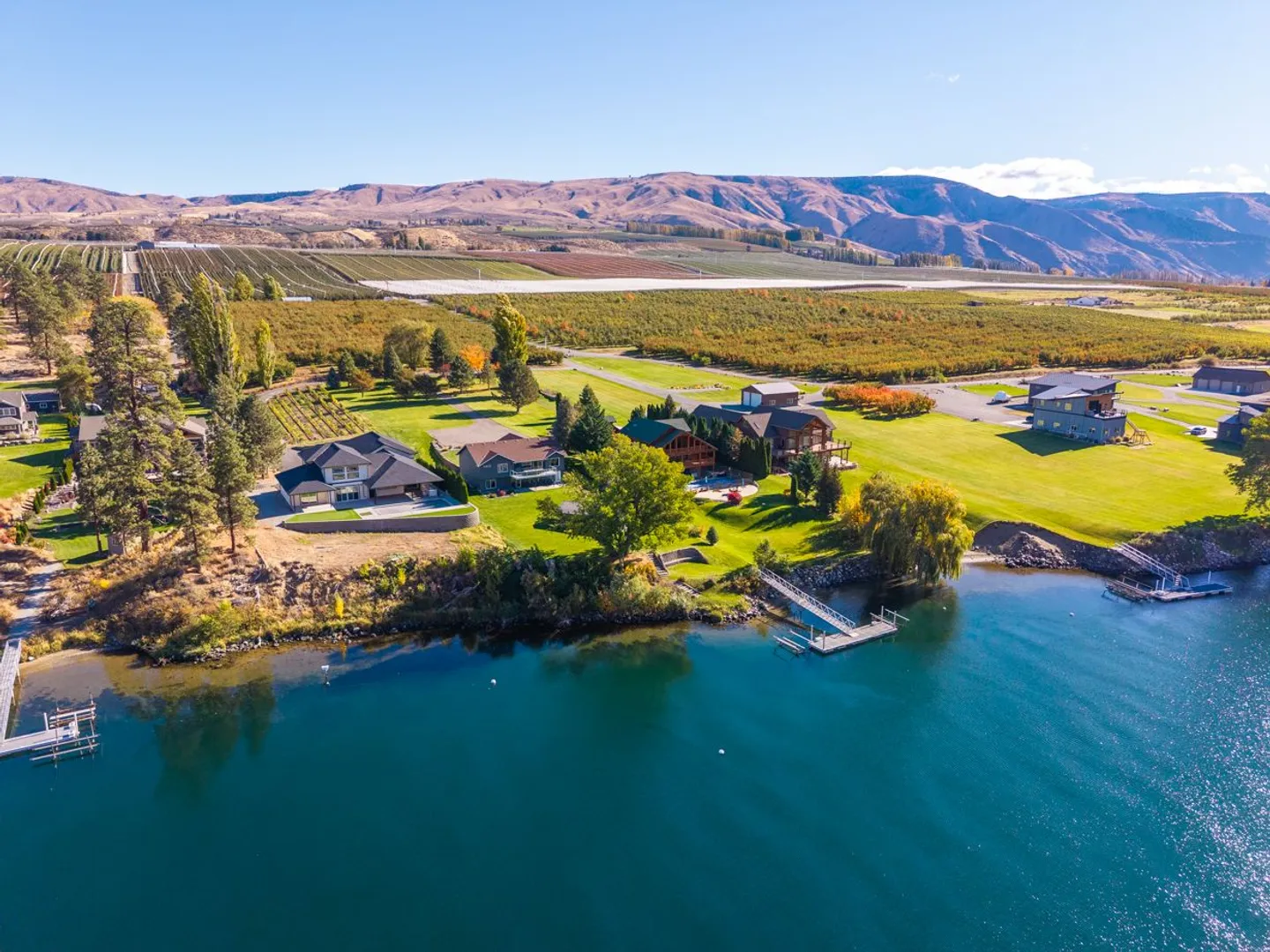 Aerial view of waterfront homes along a lake, with private docks, expansive green lawns, orchards, and arid hills in the background.