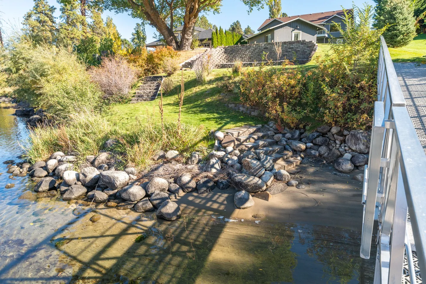 A grassy slope with stone retaining walls, rock shoreline, and stone steps leading down to the water next to a metal dock railing.