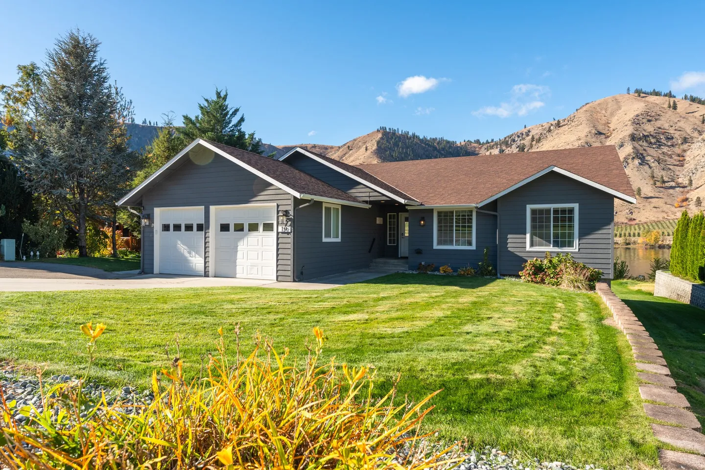 Front exterior of a single-story gray home with a two-car garage, manicured green lawn, and arid hills in the background.