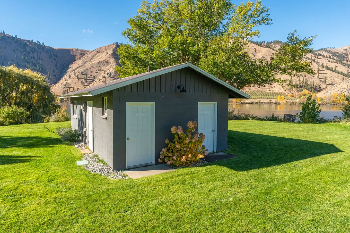 Exterior view of a dark gray outbuilding on a green lawn near a river with hills in the background.