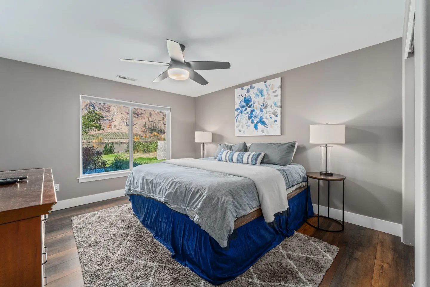 Bedroom with a large bed dressed in blue and grey linens, a picture window showing hills outside, and dark wood-style floors.