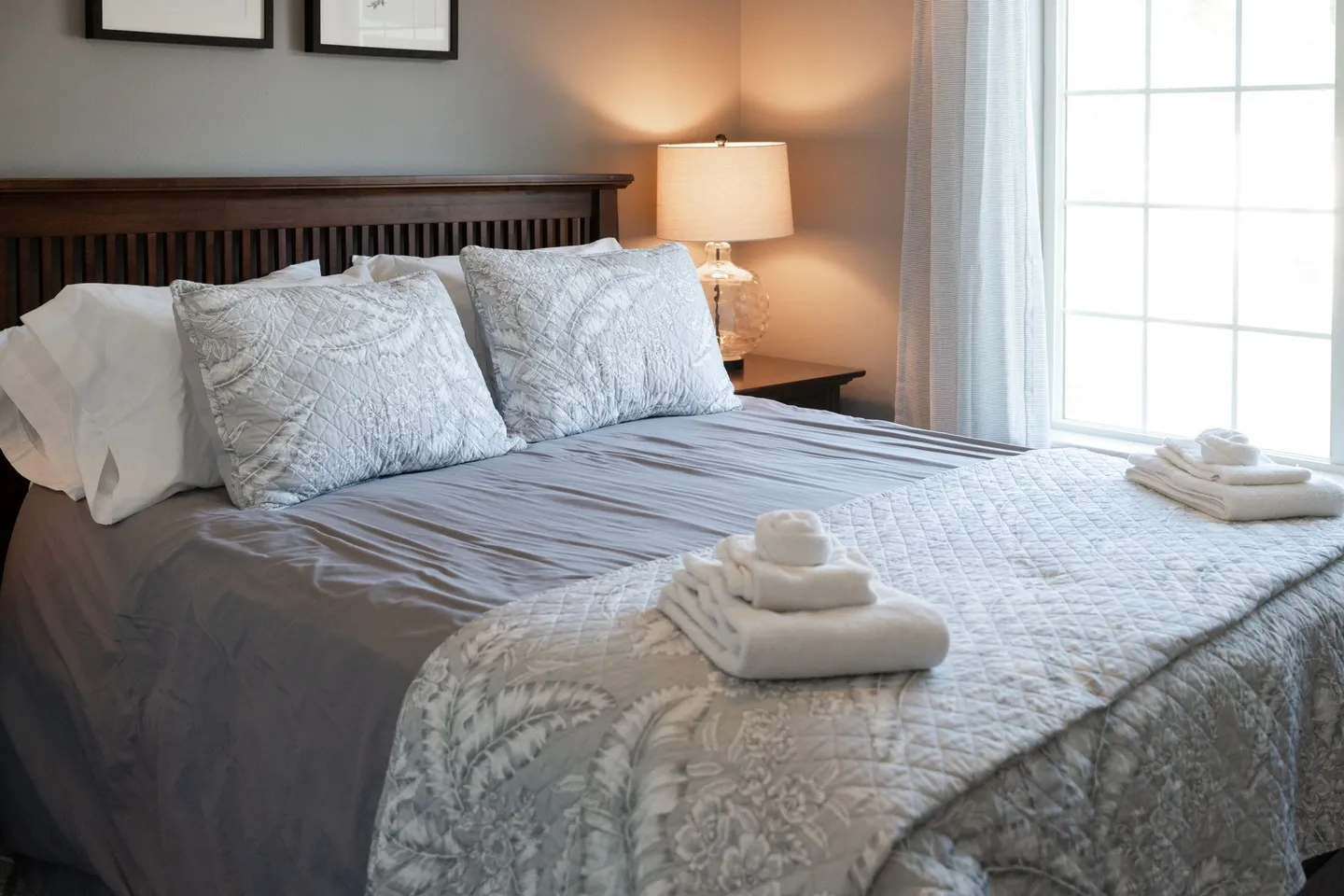 Bedroom with a wooden headboard, made bed featuring patterned grey and white bedding, folded towels, and a nightstand with a lit lamp next to a window.