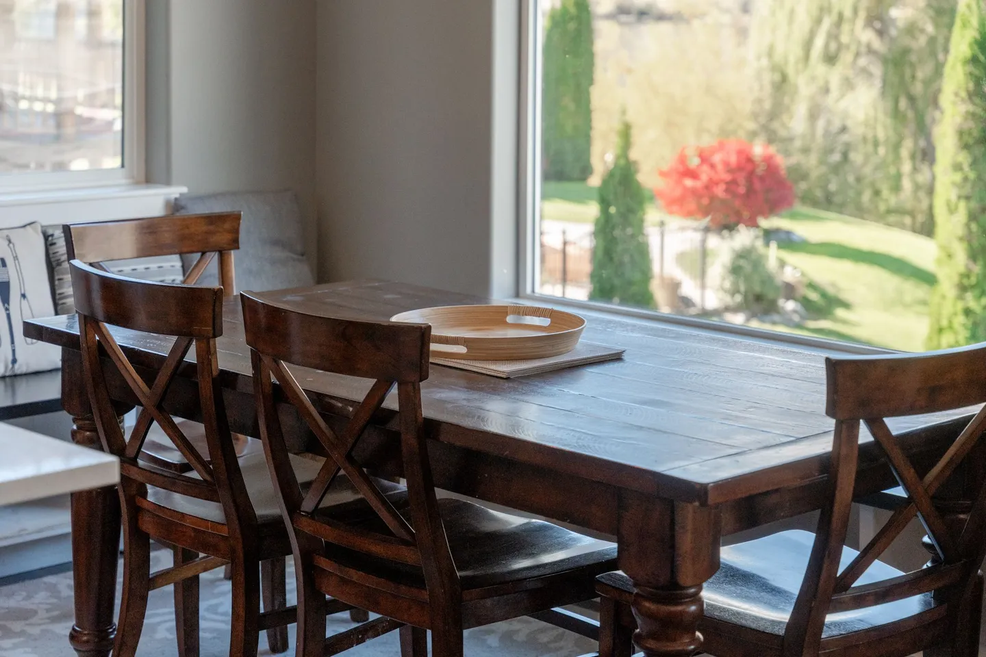 Dark wooden dining table and cross-back chairs positioned next to a large window overlooking a landscaped yard.