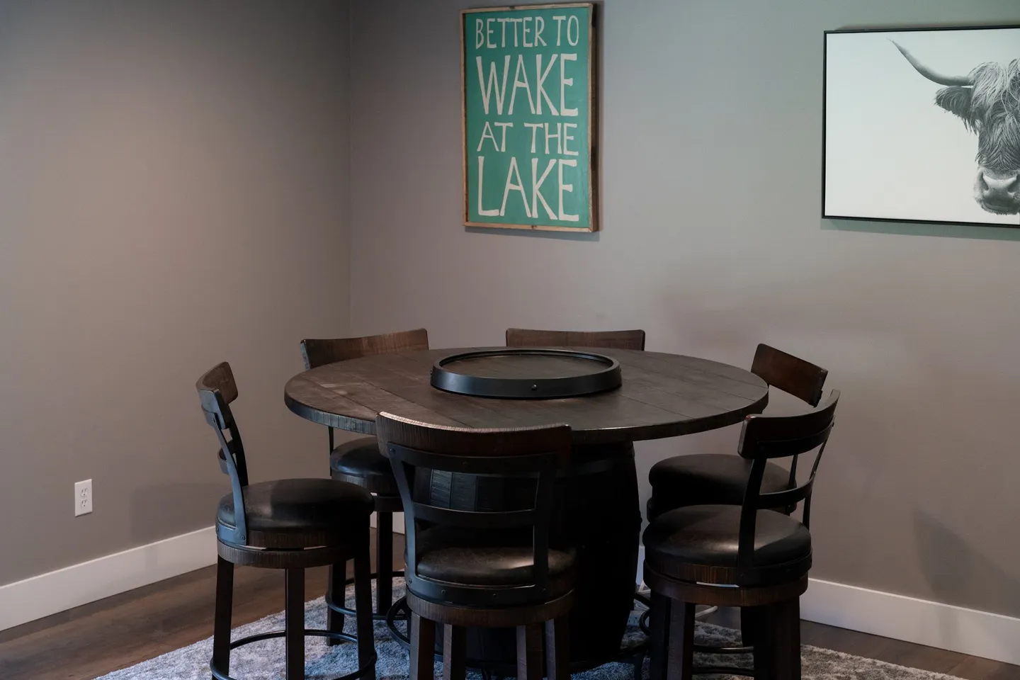 Round dark wood table with a lazy susan surrounded by barstool chairs in a room with gray walls and wall art.