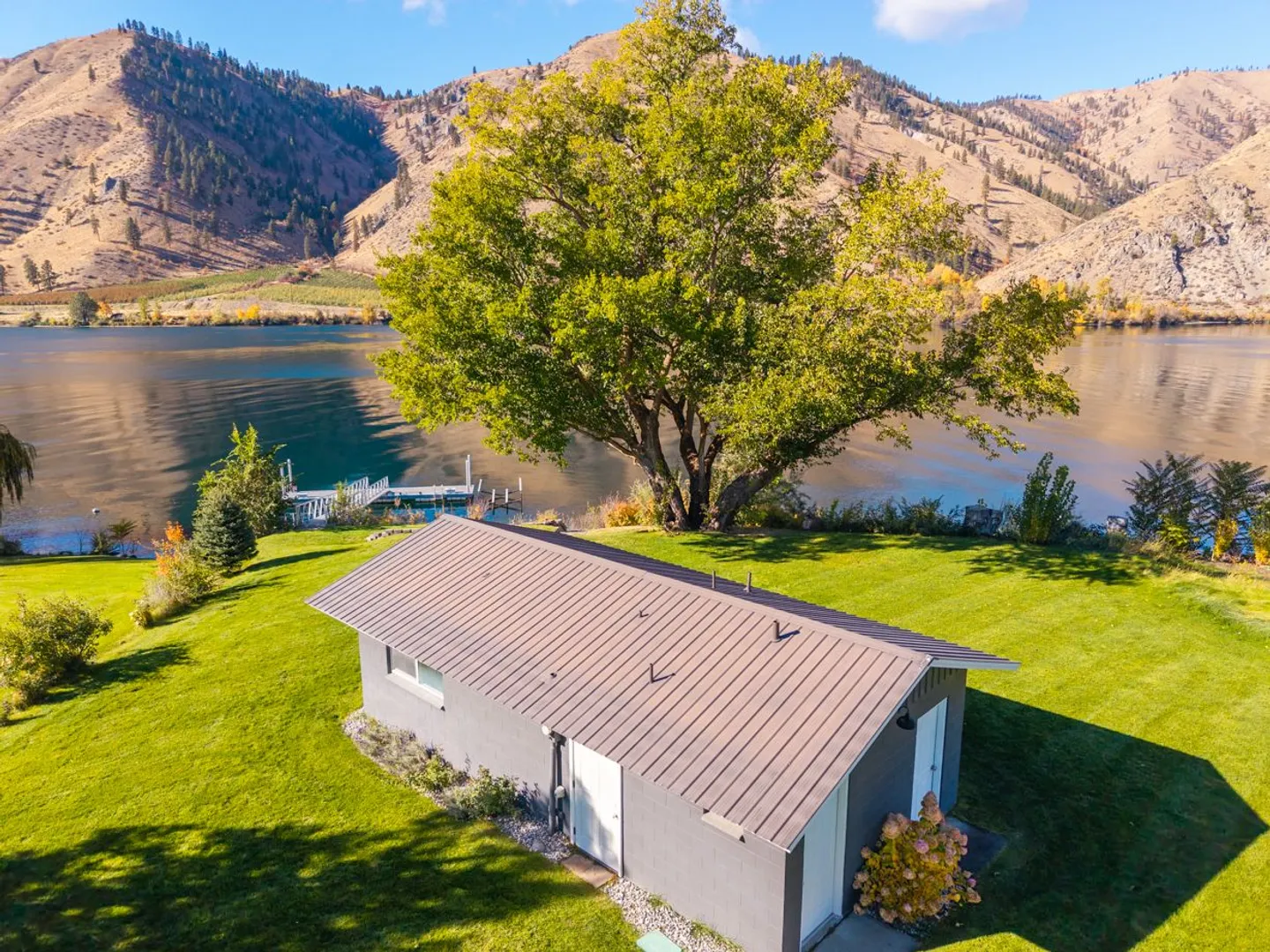 Aerial view of a gray building with a metal roof set on a green lawn beside a lake, with a dock and mountainous hills in the background.