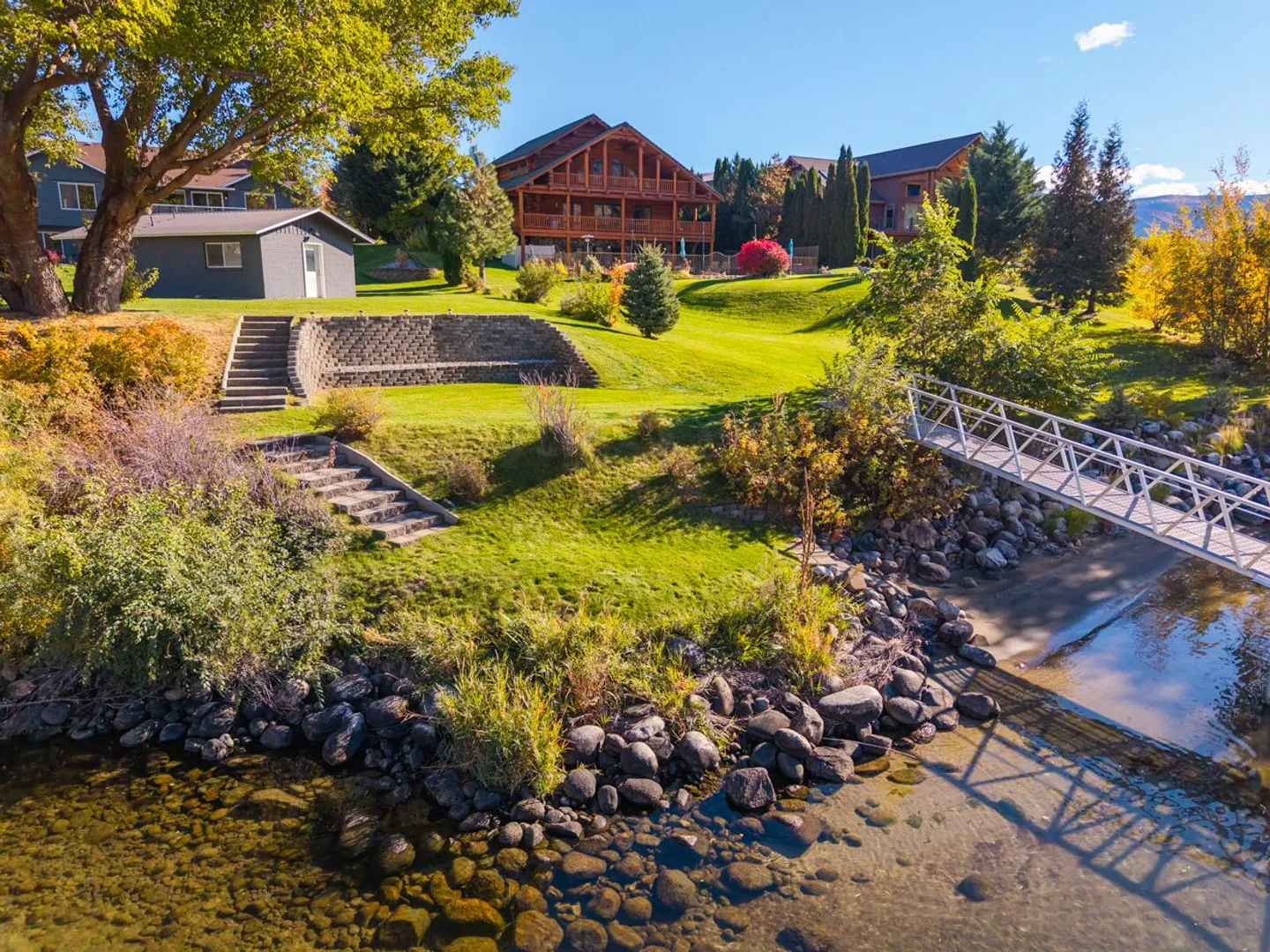 Elevated view of a waterfront property with a grassy lawn, stone steps, a footbridge to a dock, and large log homes in the background under a blue sky.