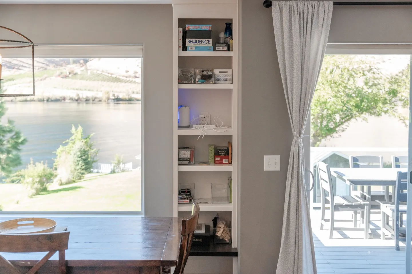 Dining area with a wooden table, built-in shelving stocked with games, a large picture window overlooking a lake, and a sliding glass door to an outdoor deck.