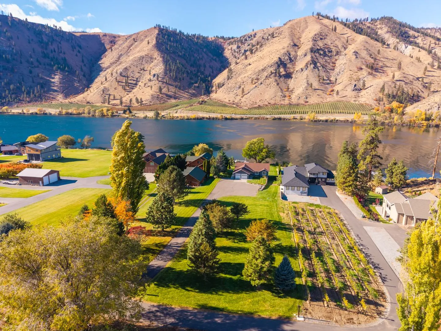 Aerial view of waterfront houses along a wide river with arid mountains rising on the opposite shore under a clear blue sky.