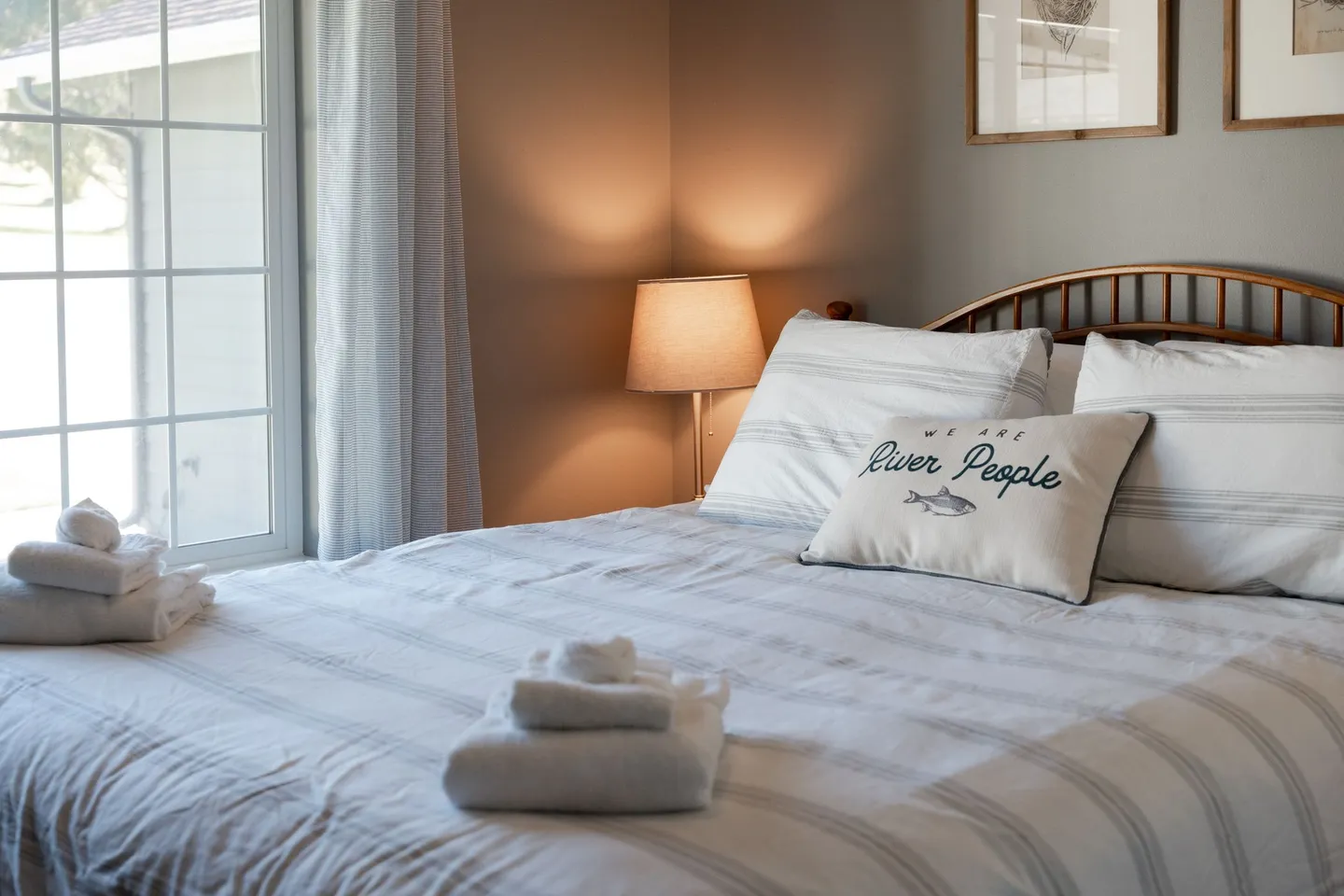 Close-up of a made bed with striped bedding, a decorative pillow reading 'We Are River People', and folded white towels.