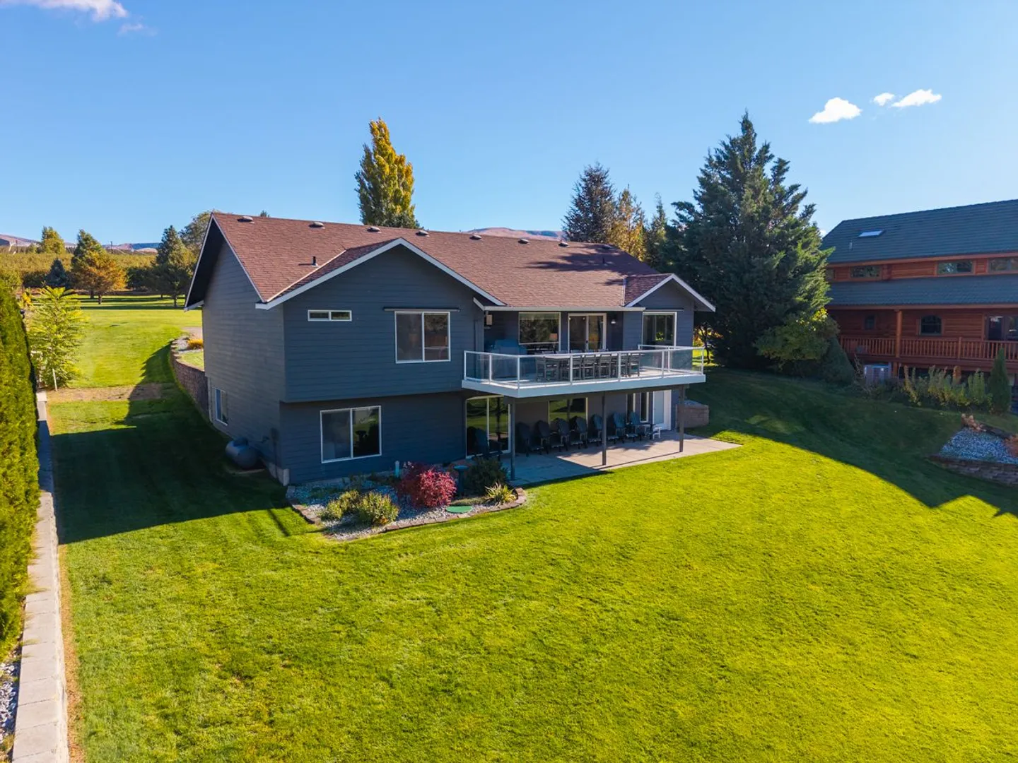 Aerial view of the rear exterior of a two-story gray house with an elevated deck, lower patio, and expansive green lawn under a blue sky.