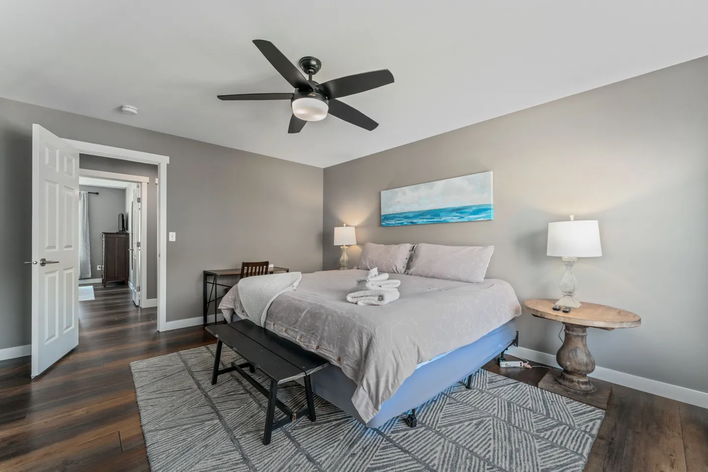 Bedroom featuring a made bed with rolled towels, wood-look flooring, ceiling fan, bedside tables with lamps, and a small desk.