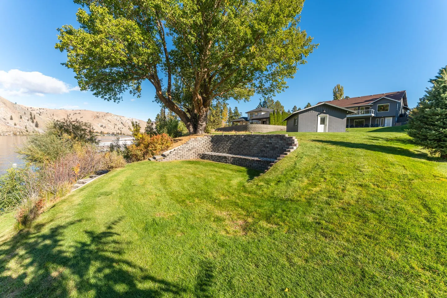 Sloping green lawn with a stone retaining wall and a large leafy tree overlooking a body of water and hills.