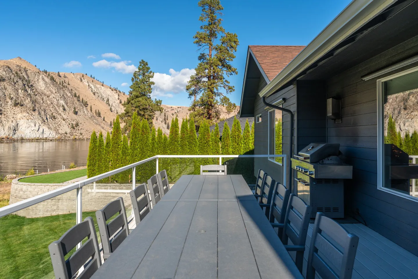 Outdoor deck with a long dining table, chairs, a barbecue grill, and views of a body of water and hills.