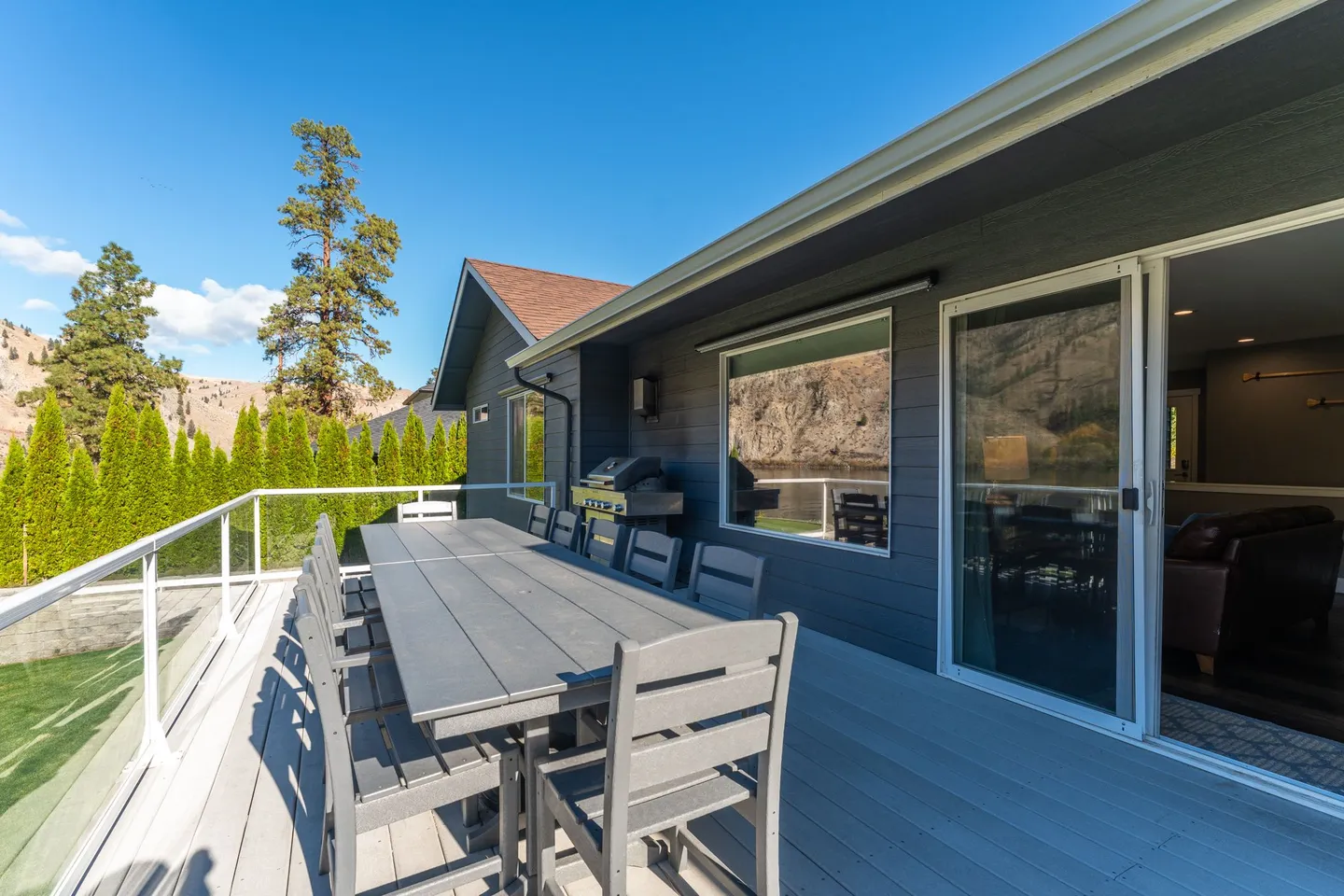 Outdoor deck with a long dining table, chairs, barbecue grill, and glass railing under a clear blue sky.