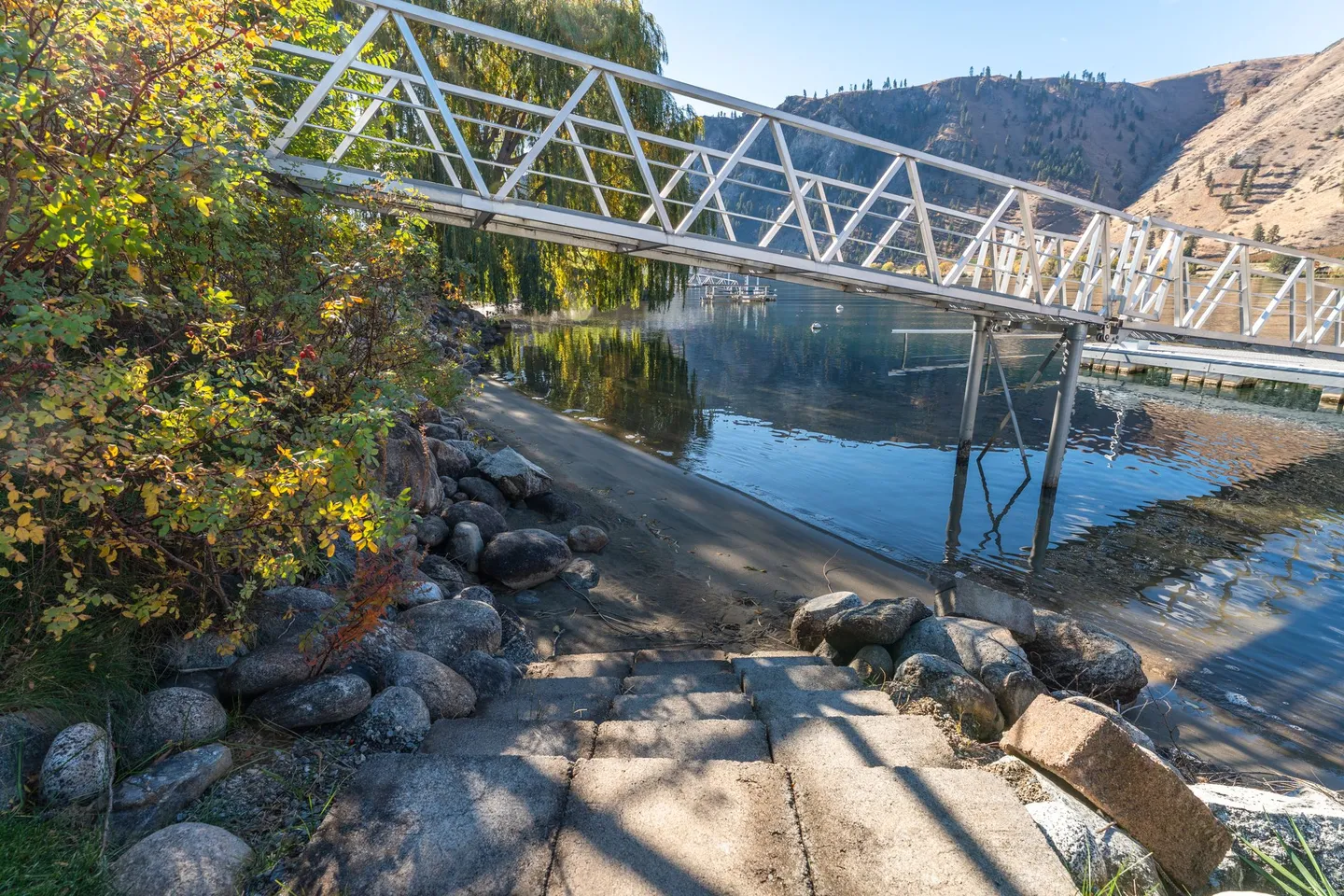 Concrete steps leading down to a sandy shoreline next to a metal dock gangway over calm water.