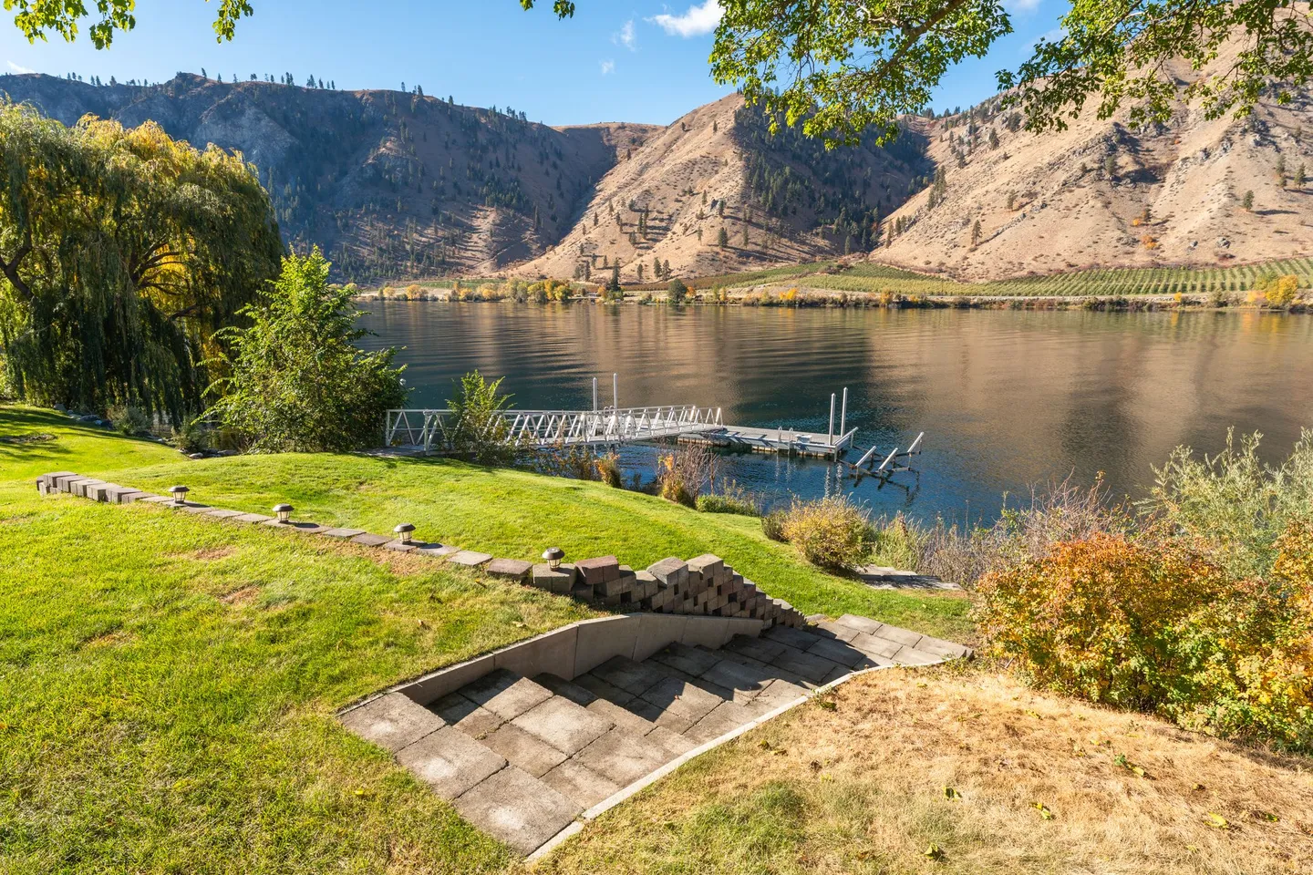Stone steps leading down a grassy slope toward a private boat dock on a wide river surrounded by arid mountains.