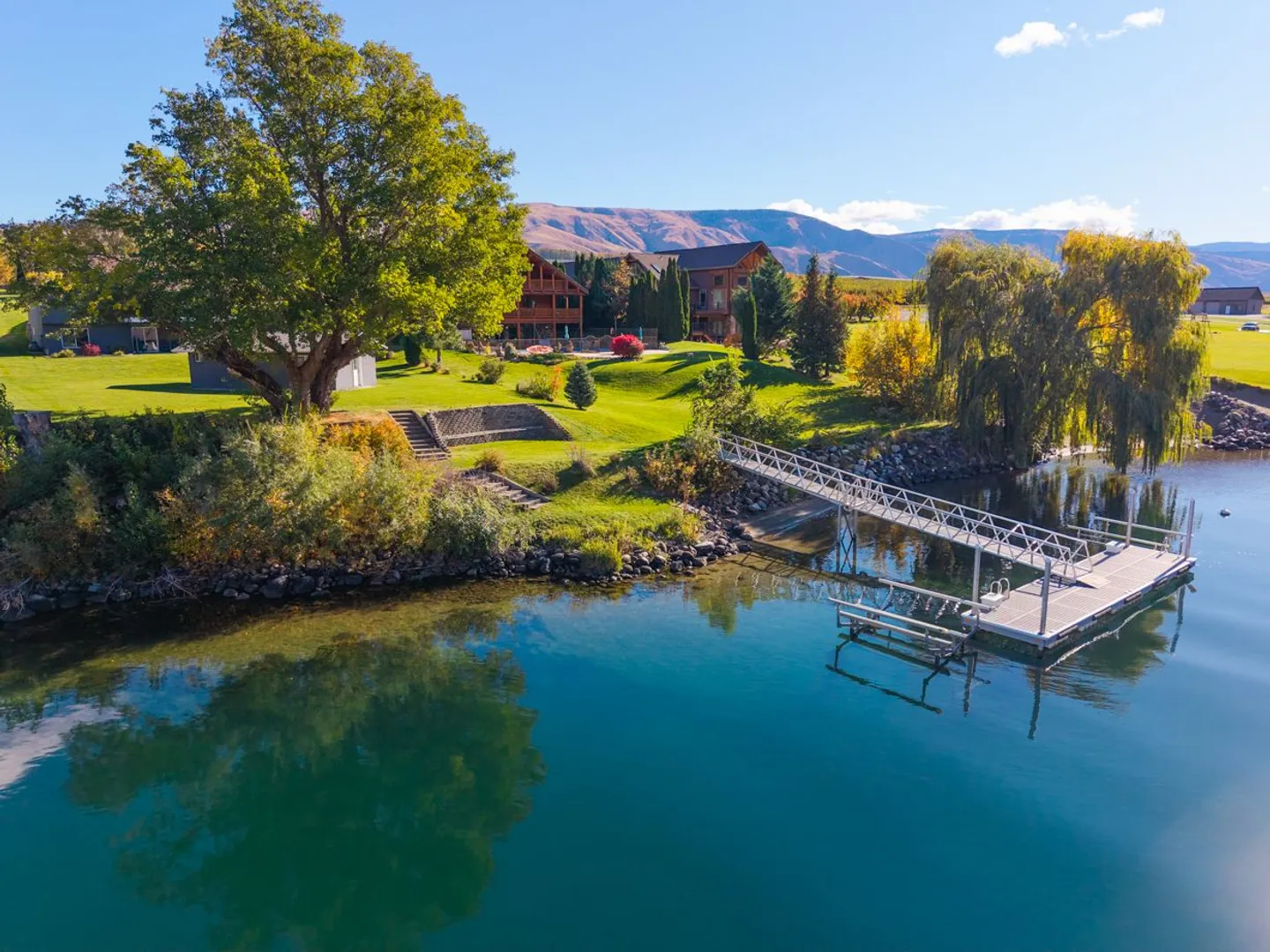 Aerial view of a waterfront property with a grassy lawn, private dock, trees, and hillside in the background.