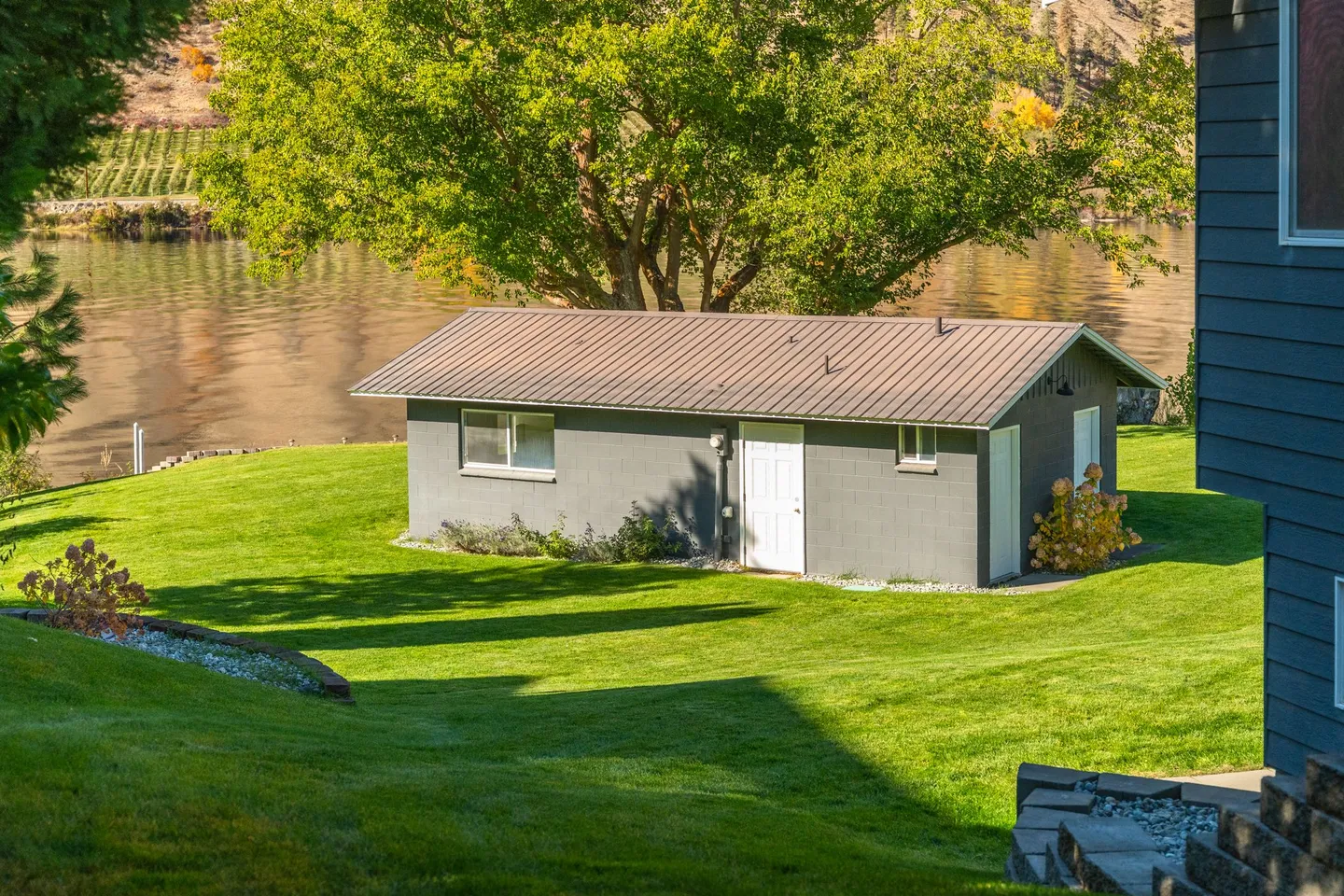 A detached grey outbuilding with a metal roof on a grassy lawn beside a body of water.