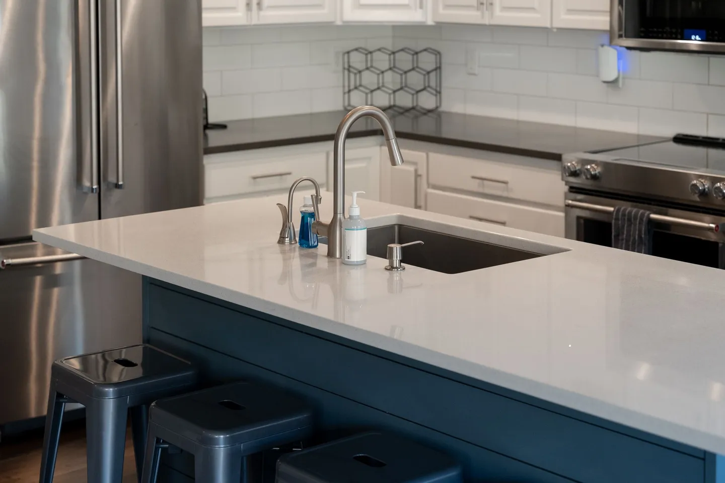 Close-up view of a kitchen island with an undermount sink, goose-neck faucet, and bar stools, with stainless steel appliances in the background.