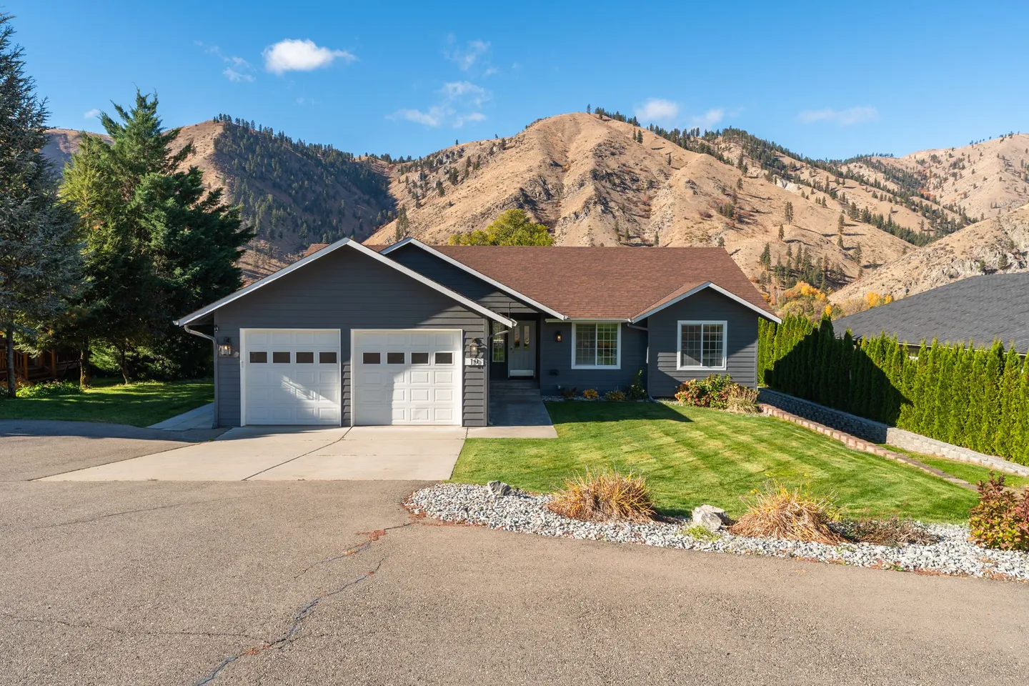 Front exterior of a single-story dark gray house with a two-car garage, manicured lawn, and arid mountains in the background.