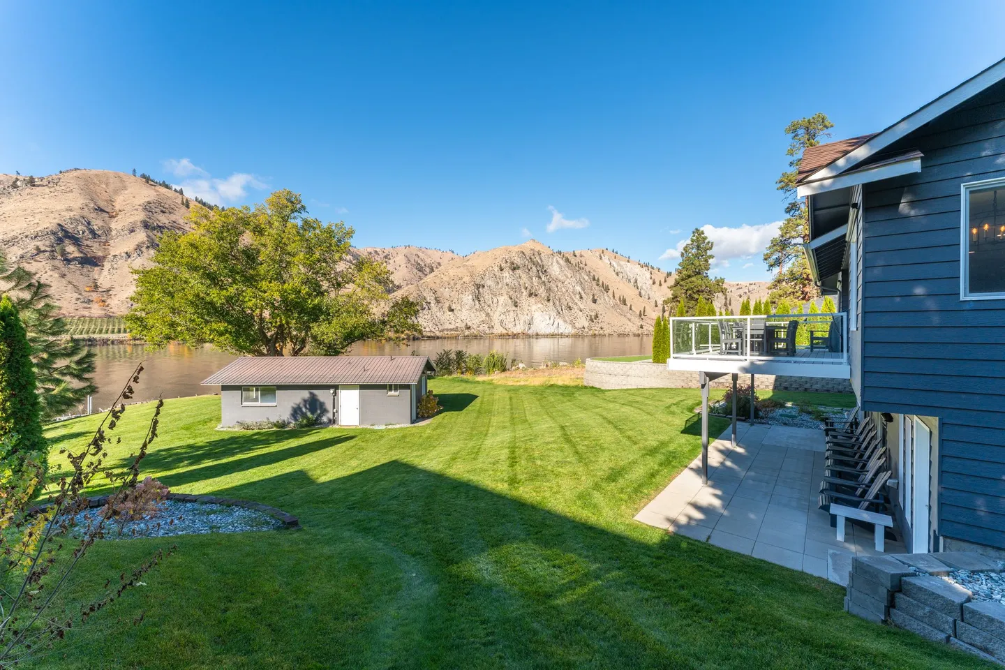 Wide view of a sloping green lawn behind a blue multi-level home, overlooking a body of water and mountains.