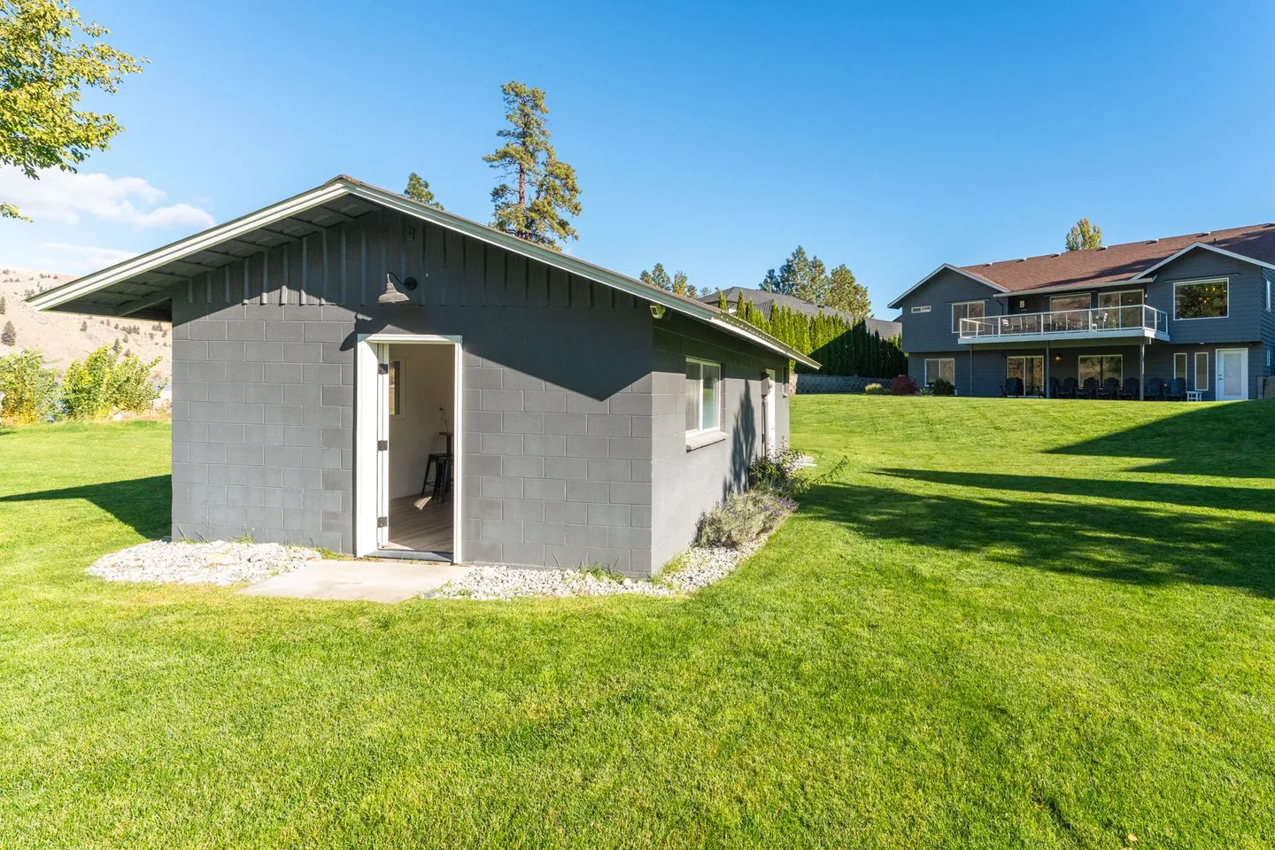 Exterior view of a gray outbuilding on a green lawn with a two-story house visible in the background under a blue sky.