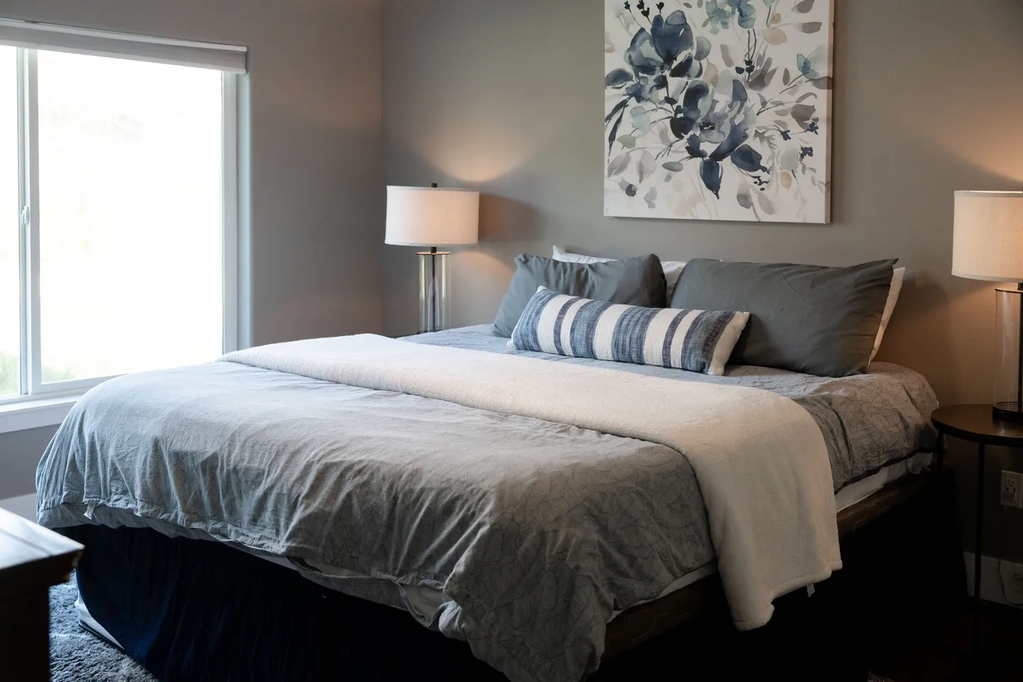 Bedroom featuring a made bed with grey and white linens, flanked by matching nightstands with lamps and floral wall art above.