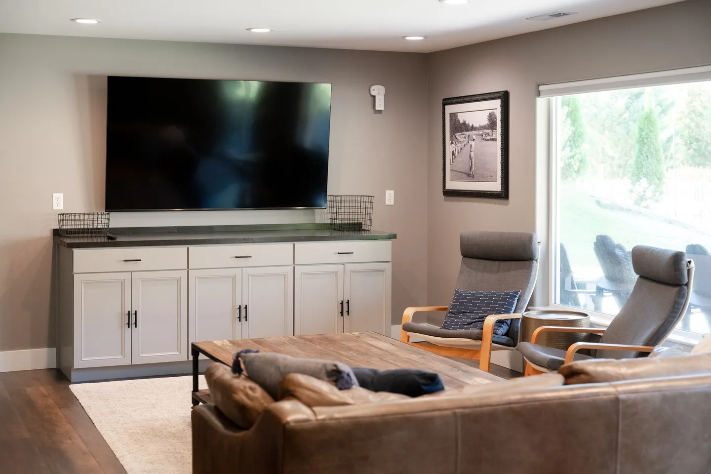 Living room with a large wall-mounted flat-screen TV above gray cabinetry, a leather sofa, two armchairs, and a sliding glass door.