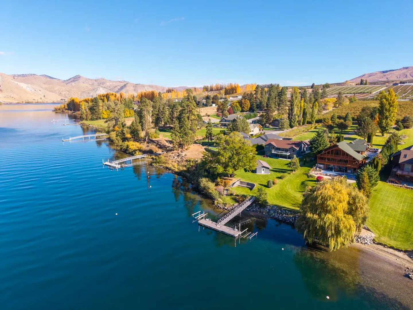 Aerial view of a lakefront residential community with private docks, green lawns, and autumn trees against rolling hills and a clear blue sky.