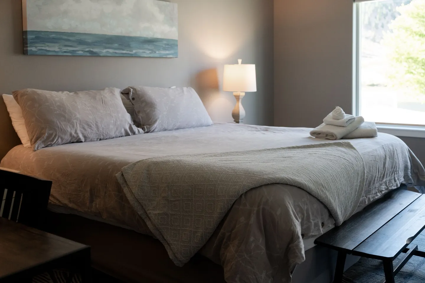 Bedroom with a made bed, folded towels, a bedside lamp, ocean artwork on the wall, and a window.