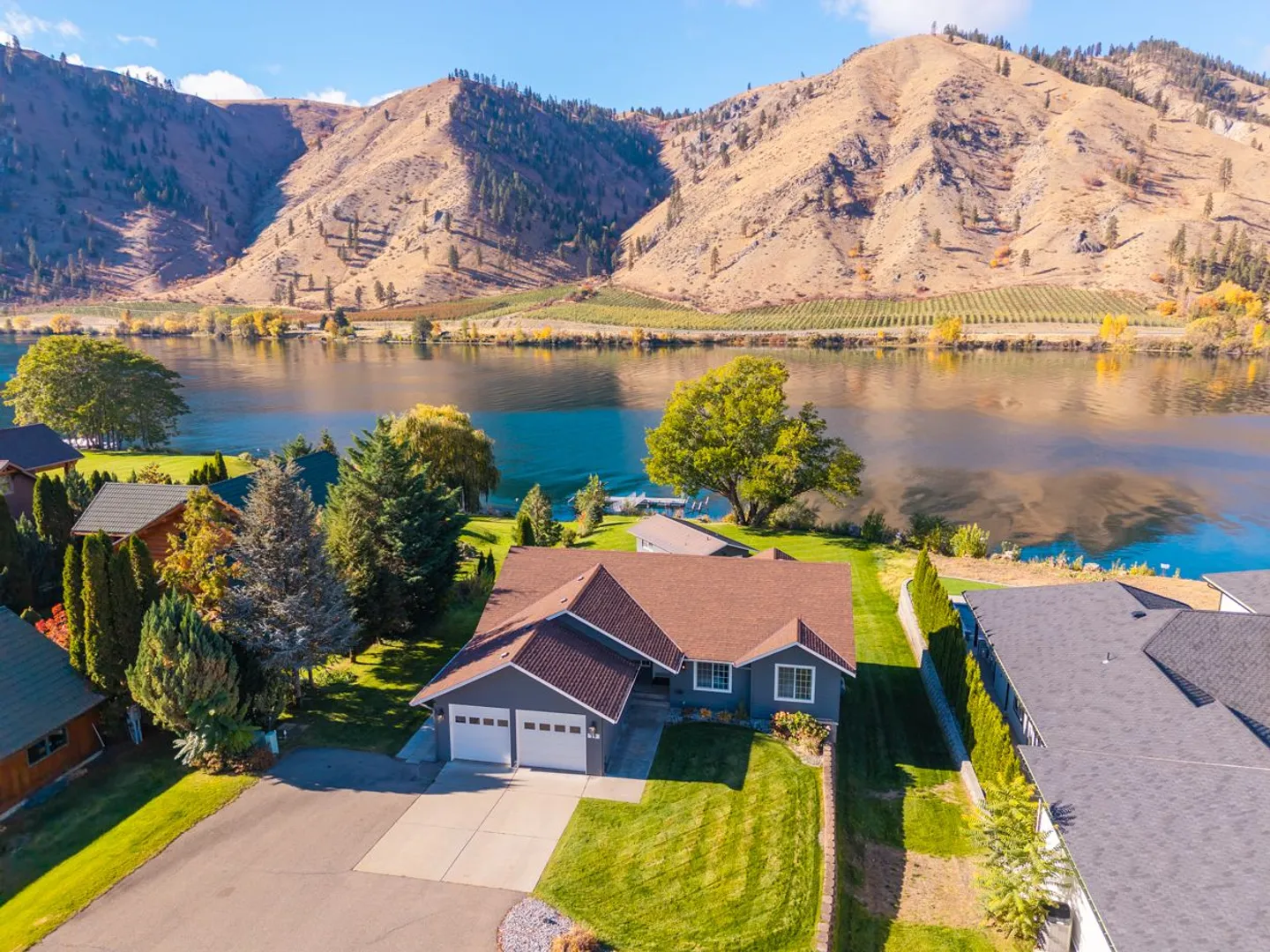 Aerial view of a single-story waterfront home with a two-car garage, front lawn, and views of a wide river and mountains.