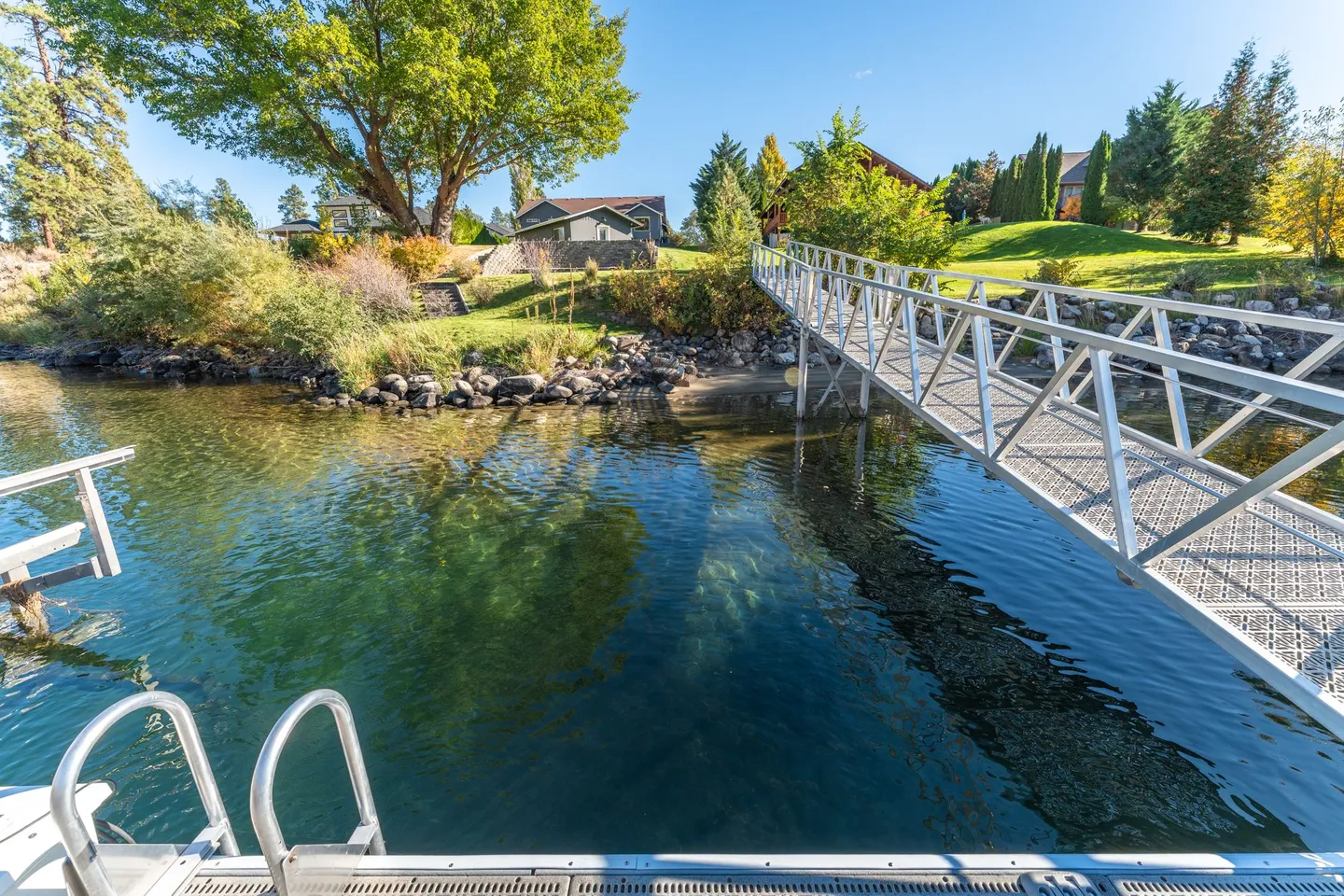 View from a boat dock looking across clear water toward a metal gangway leading to a grassy shoreline with houses.