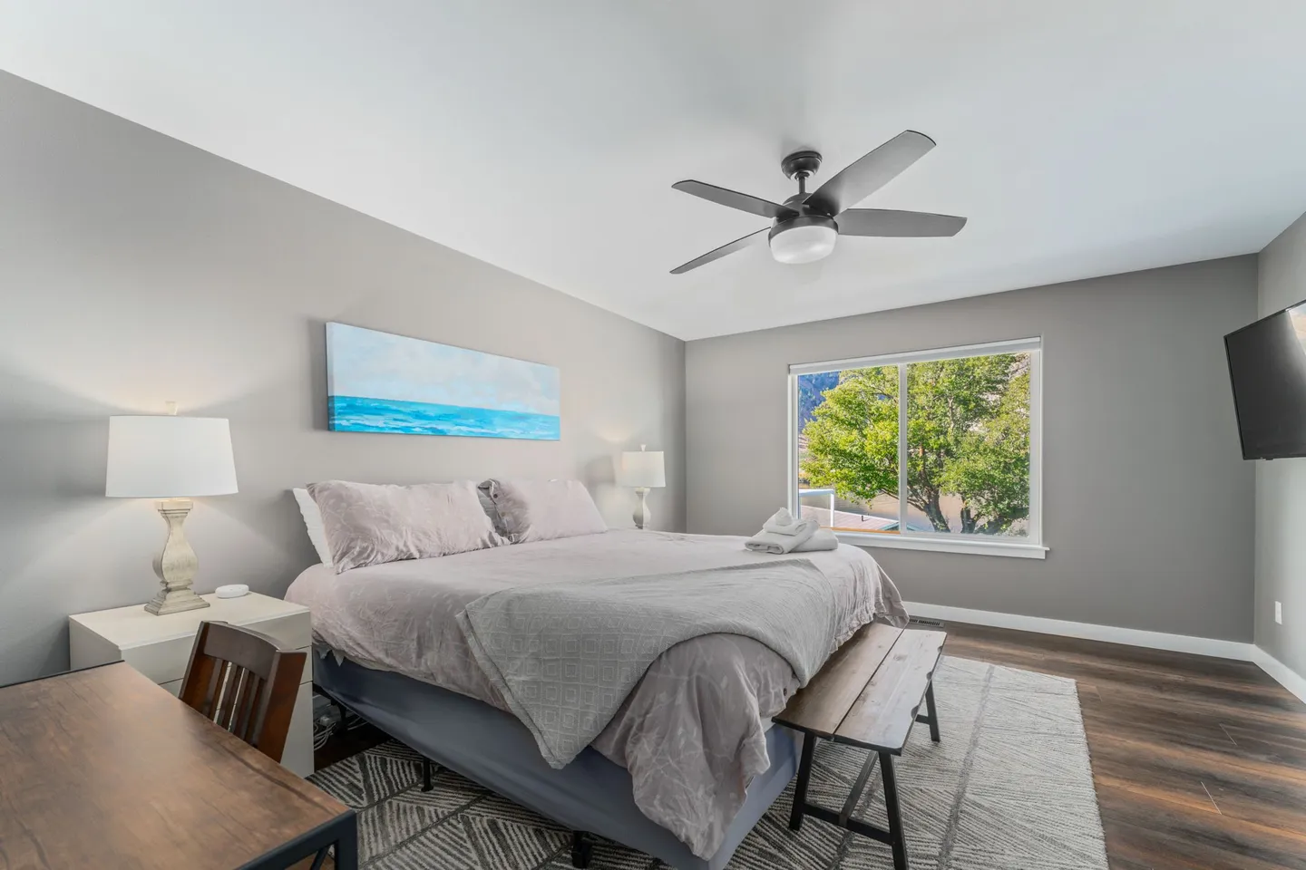 Bedroom with a large bed, nightstands with lamps, a ceiling fan, and a window overlooking green trees.