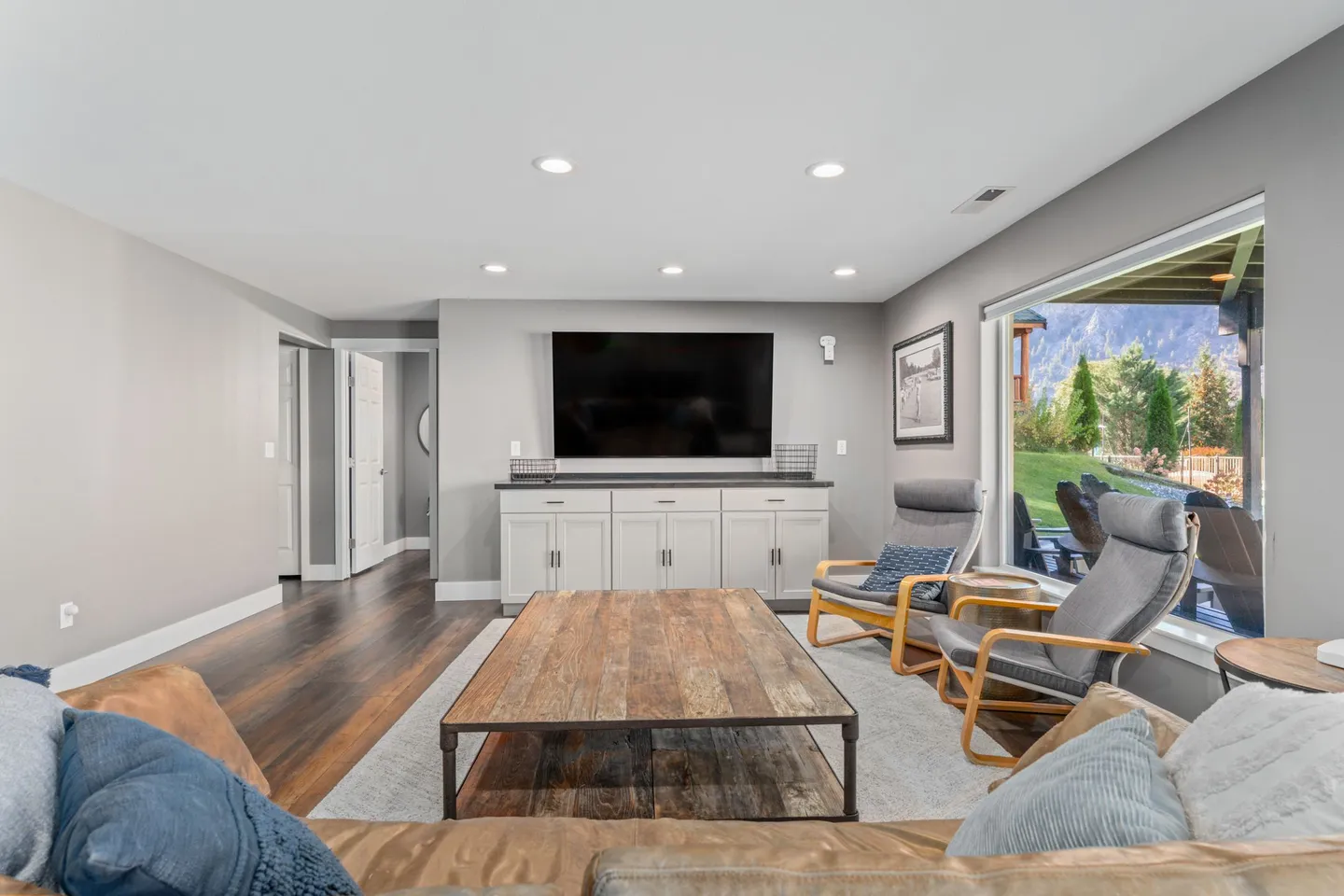 Living room with a large TV mounted over a white cabinet, a wood coffee table, armchairs, and a view of mountains through a glass sliding door.
