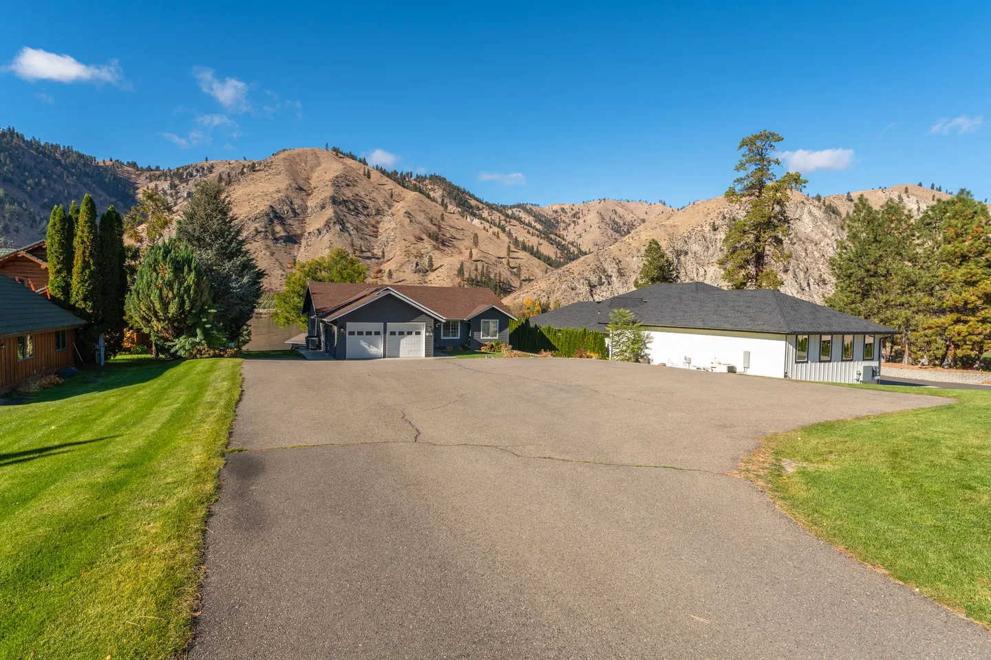 Wide asphalt driveway leading toward two residential homes backed by arid mountains under a blue sky.