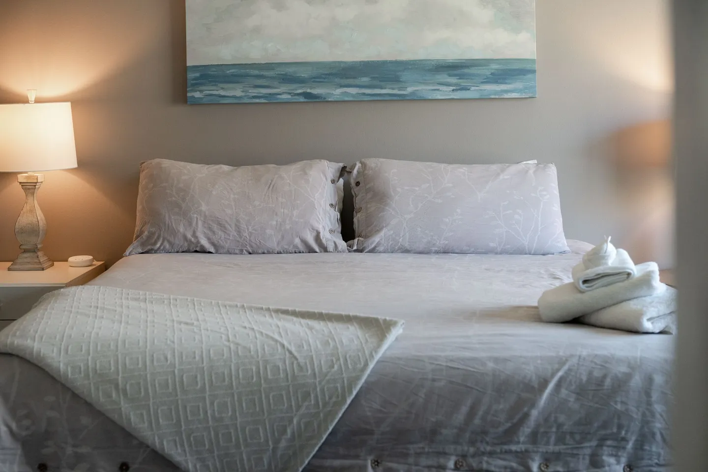 A made bed with pillows, folded white towels, a bedside table with an illuminated lamp, and an ocean painting on the wall.