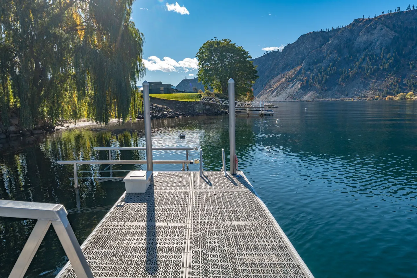 A floating boat dock extending into a clear lake with hills and trees along the shoreline.
