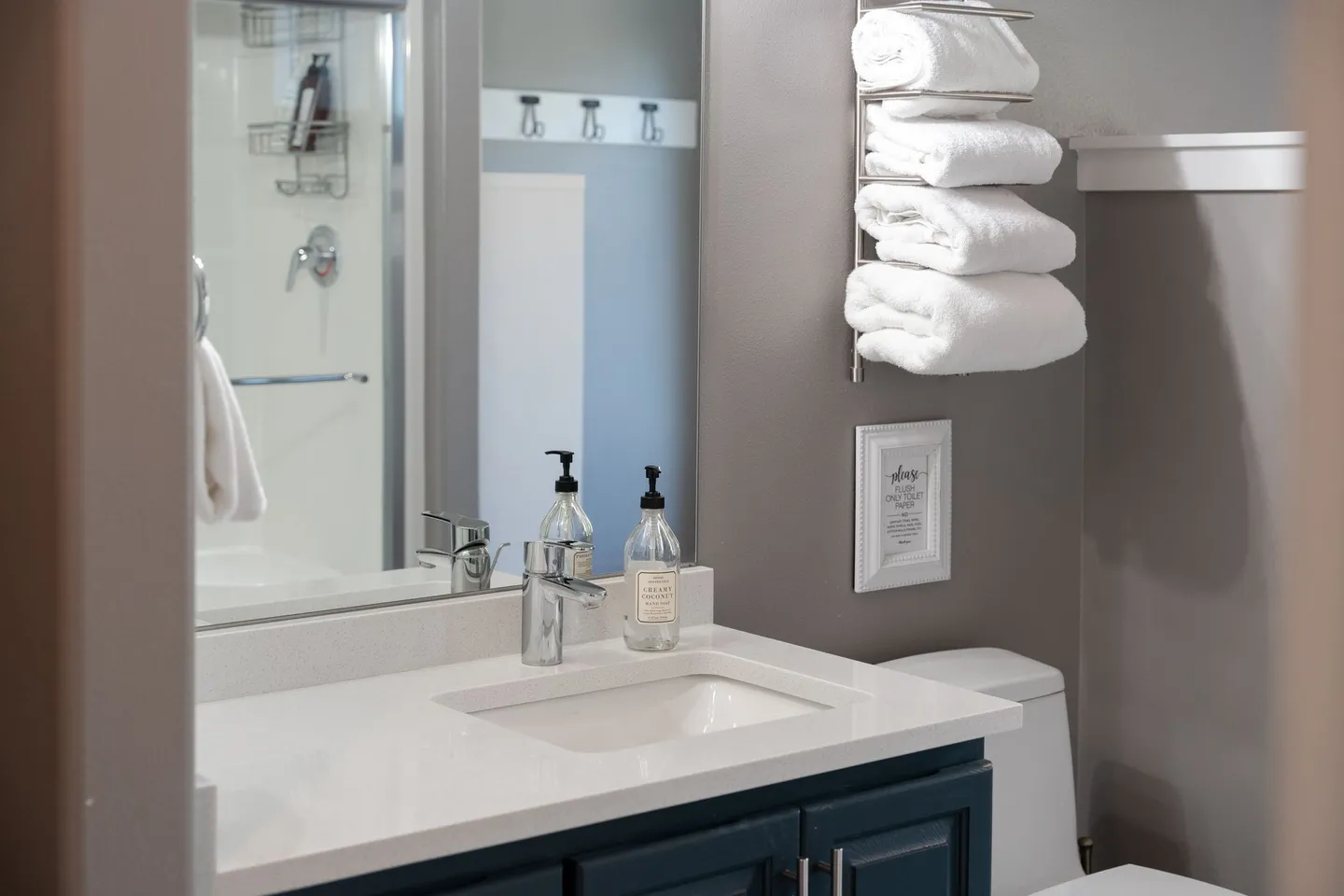 Bathroom vanity with a white countertop, modern chrome faucet, soap dispensers, mirror reflection of the shower, and stacked rolled towels on a wall rack.