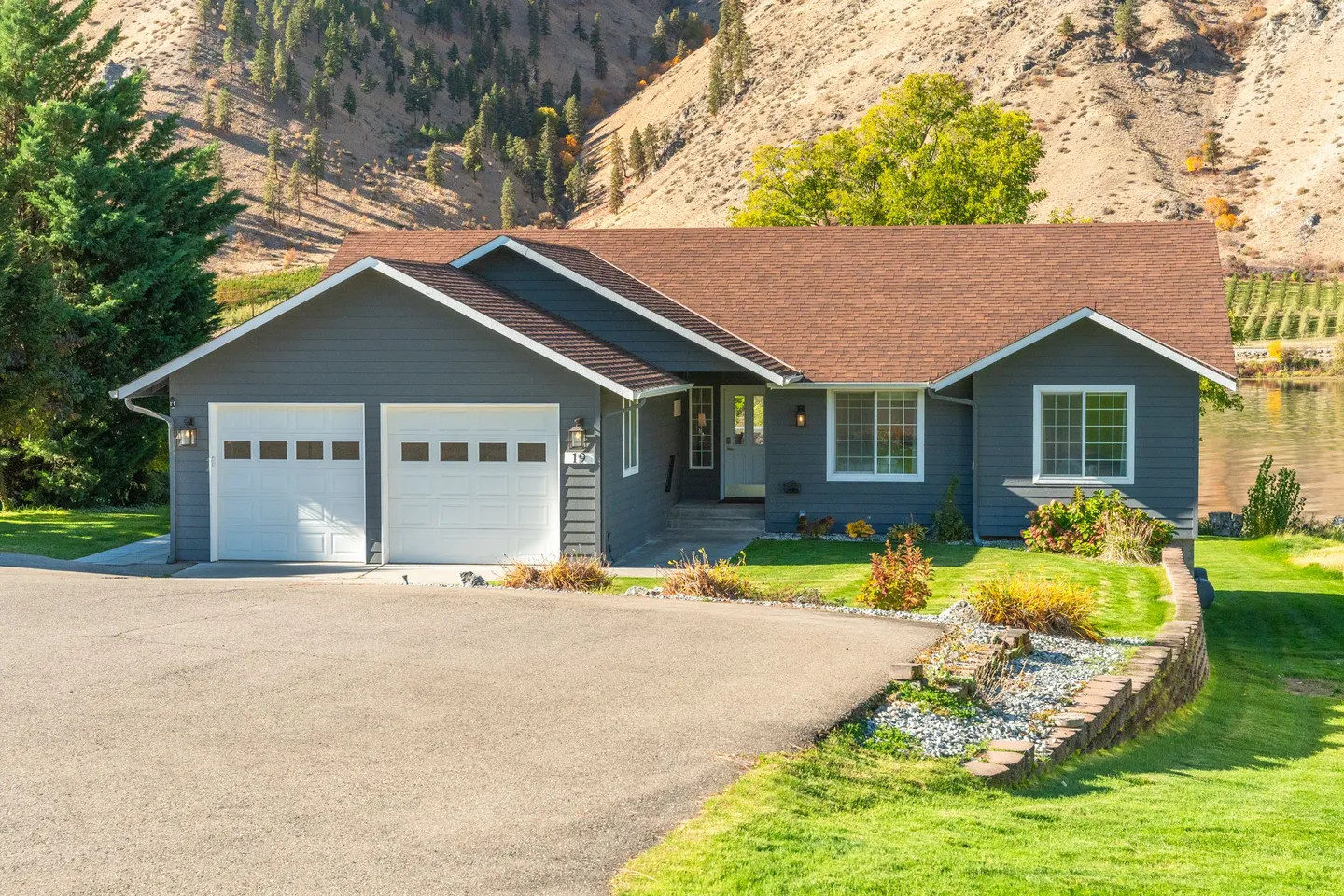 Exterior front view of a grey single-story house with a two-car garage and paved driveway, set against a hillside and water.
