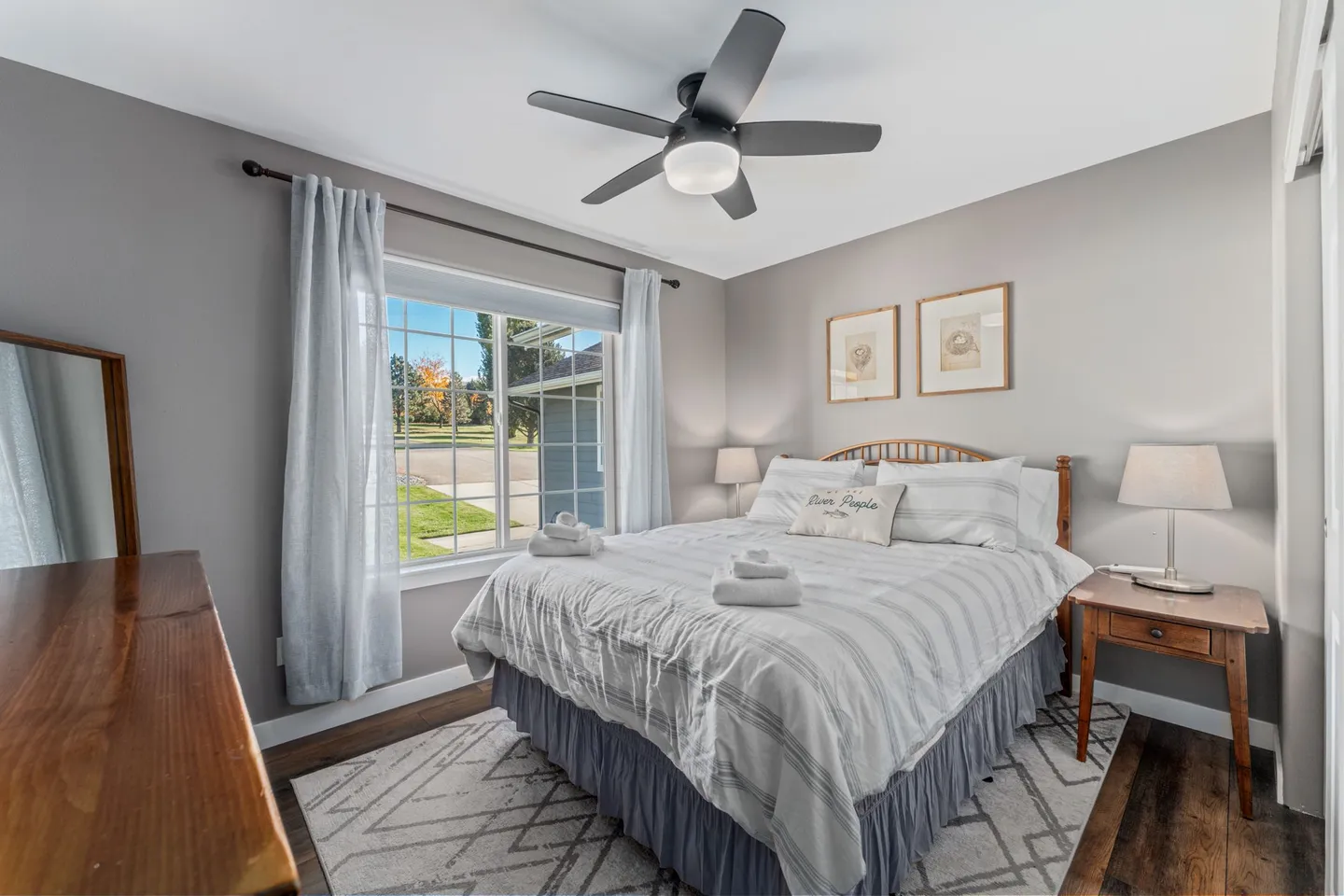 Bedroom featuring a made bed with white and gray bedding, two nightstands with lamps, a window with curtains, and a ceiling fan.
