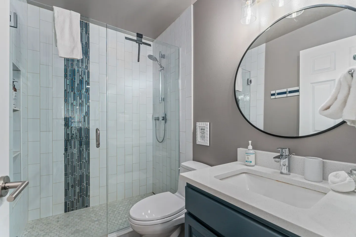 Bathroom with a glass-enclosed walk-in shower featuring blue accent tile, a white toilet, and a vanity with a circular mirror.