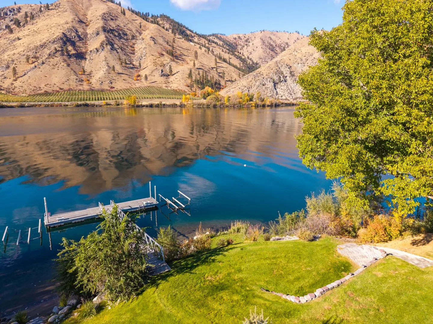 Elevated view of a calm lake and private dock bordered by a green lawn and rugged mountain hillsides.