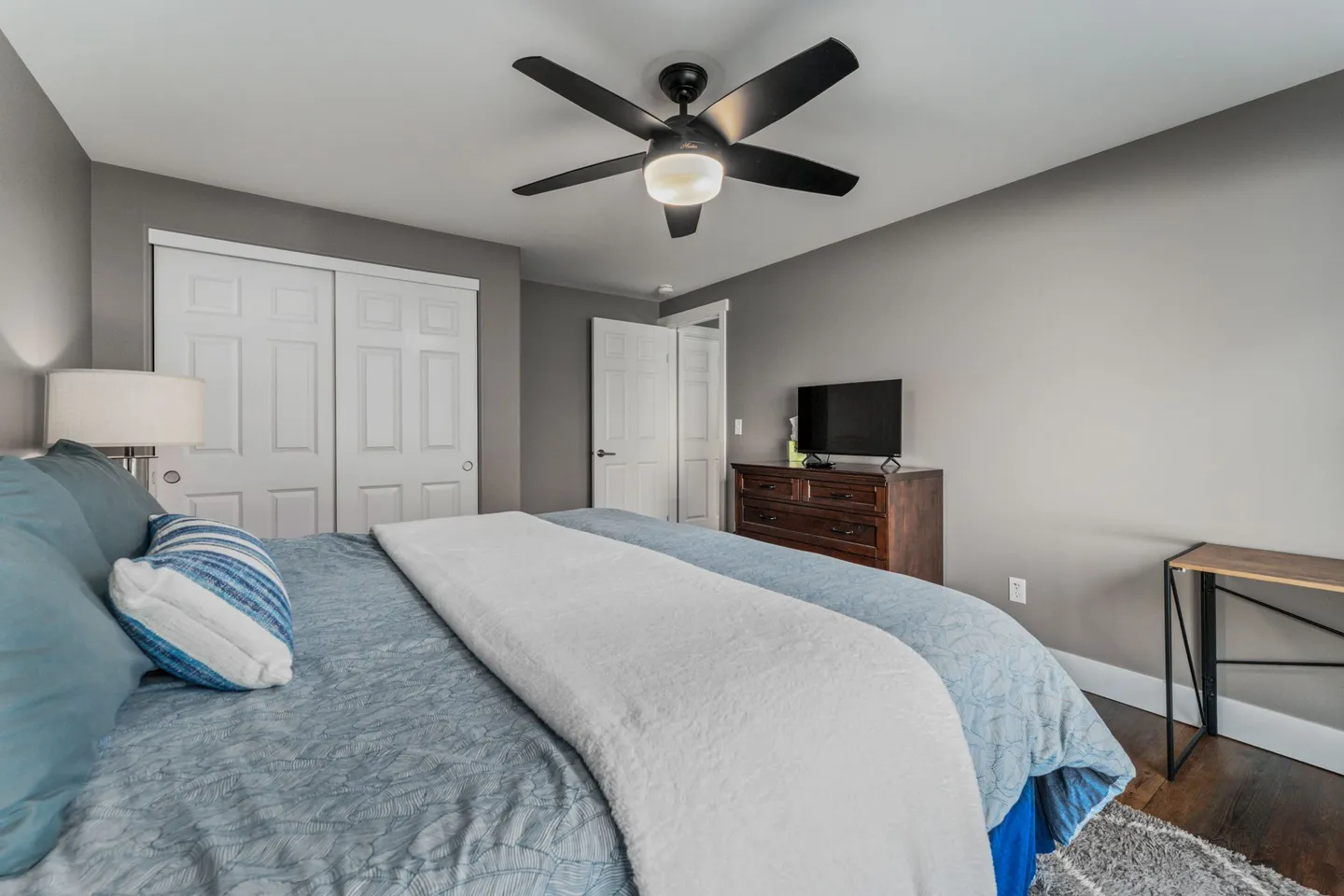 Bedroom with a bed made in blue and white bedding, a ceiling fan, closet doors, and a TV on a wooden dresser.