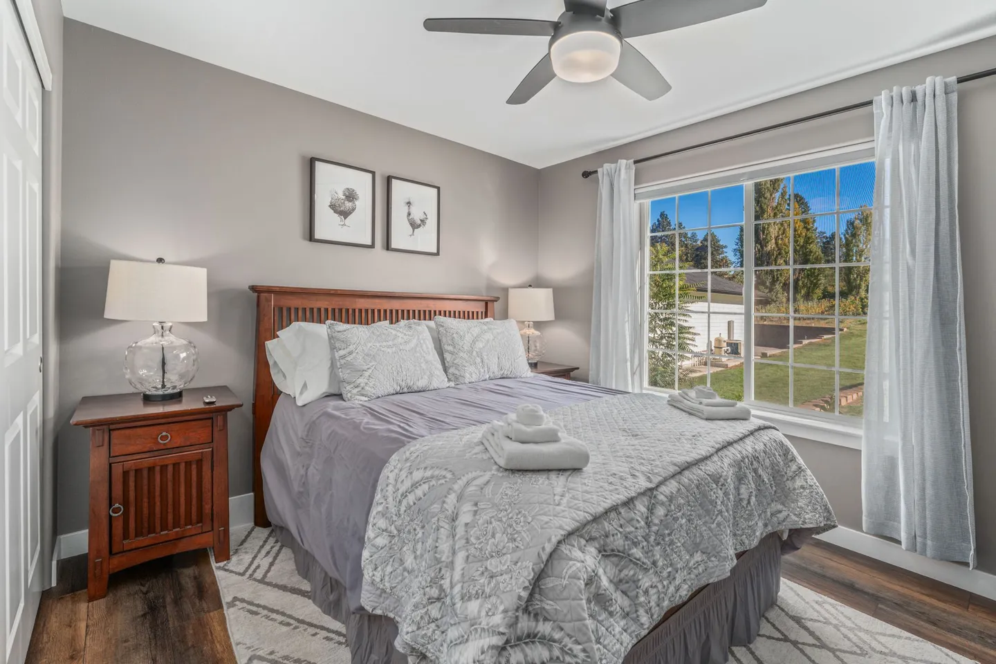 Bedroom with a wood-frame bed, gray bedding, a ceiling fan, and a large window looking out onto the yard.
