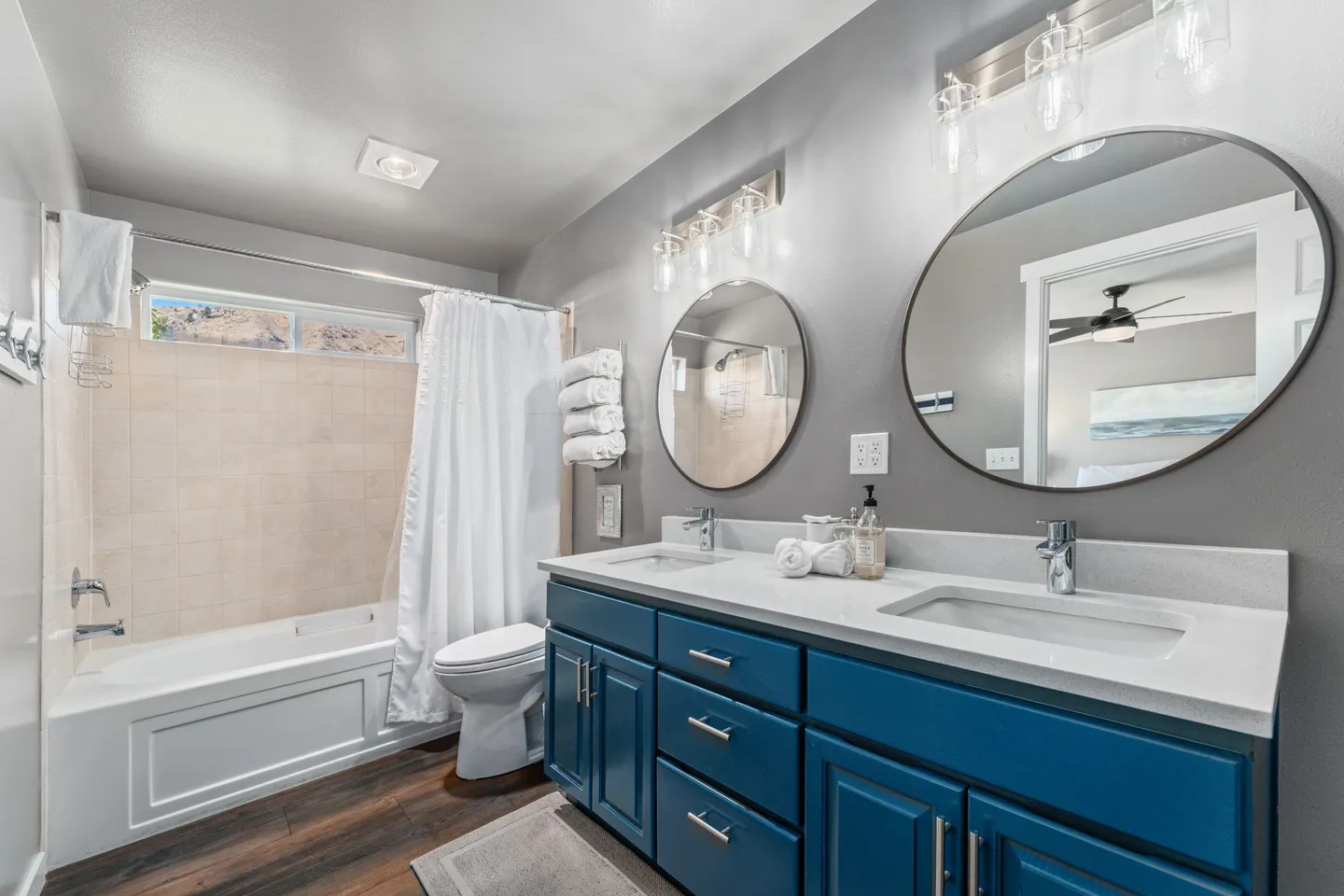Bathroom featuring a double vanity with blue cabinetry, two round mirrors, a toilet, and a bathtub with tile surround and a window above.